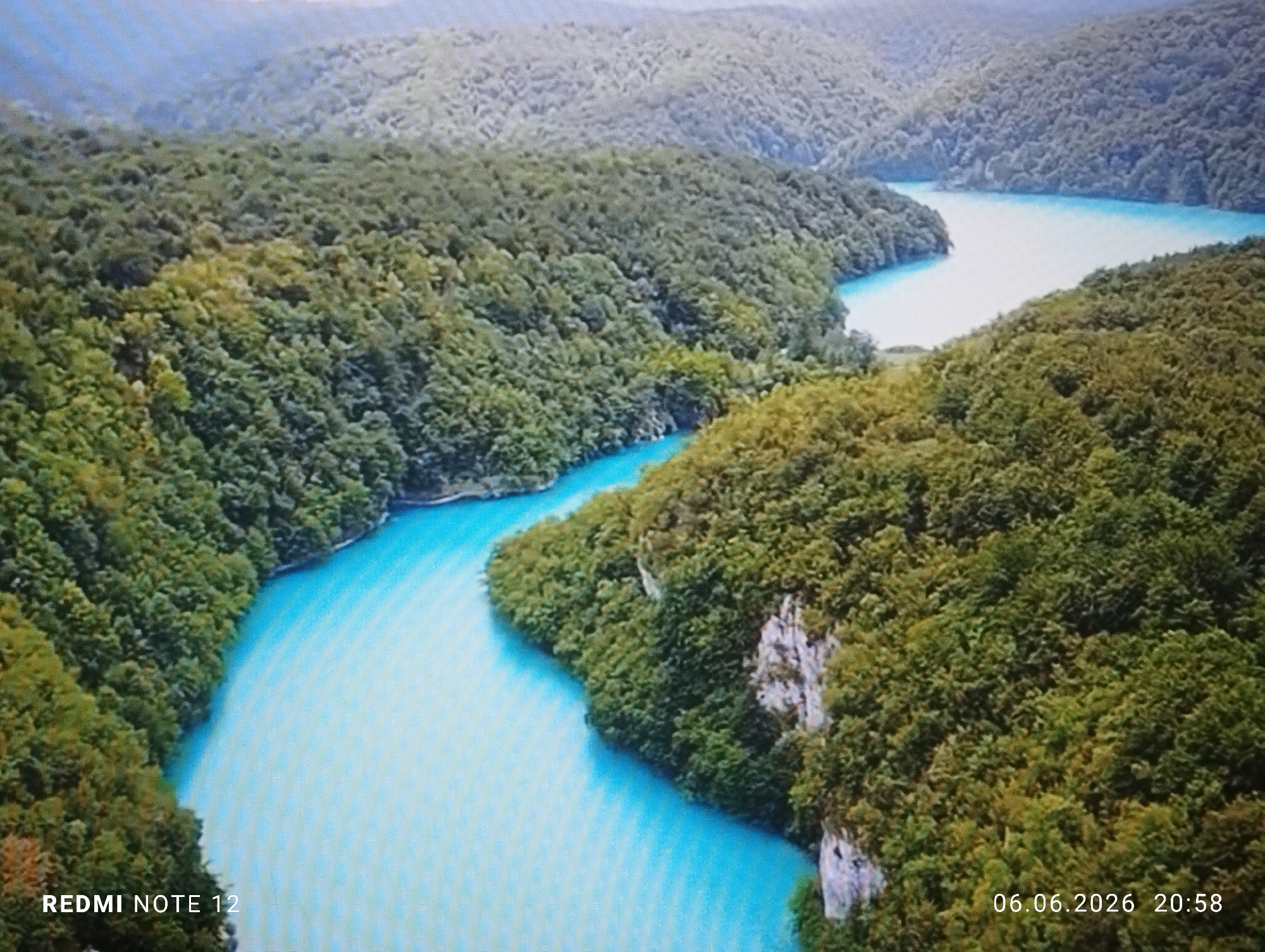 An aerial or high-angle shot depicts a pristine natural landscape dominated by a winding river or series of interconnected lakes featuring strikingly vibrant turquoise-blue water. The water body snakes through a deep valley, bordered on both sides by steep, densely forested mountains and hills. The forests consist of various shades of green, indicating a mix of tree species, with some lighter green foliage potentially suggesting new growth or specific tree types. Patches of light-colored rock or small cliff faces are visible intermittently among the trees along the water's edge, particularly on the right side of the image. The distant mountains appear softened by atmospheric haze under a light, hazy sky. No human figures, man-made structures, or vehicles are present within the natural scene. The image displays a distinct pixel grid pattern, consistent with a photograph taken of a digital screen. Overlaid text appears in the lower-left corner reading "REDMI NOTE 12" and in the lower-right corner displaying "06.06.2026 20:58". The geographical features of this image, characterized by mountainous forests and distinctively colored karst lakes, are not present in the city of Burgas, Bulgaria.