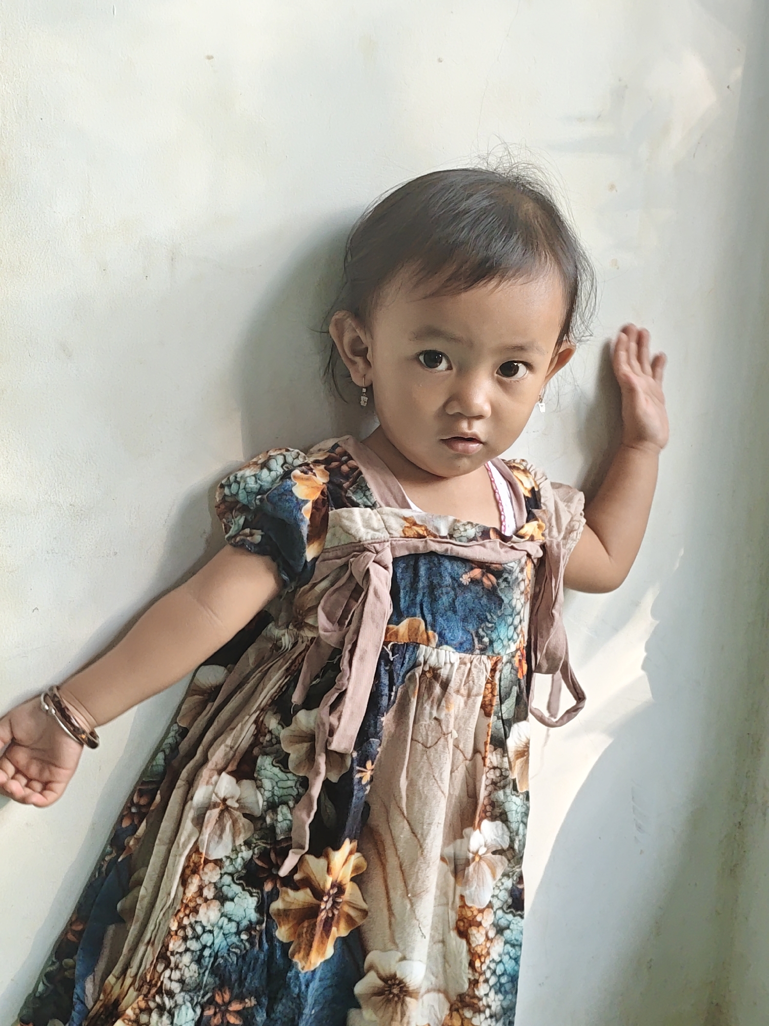 A young girl with dark hair and eyes stands against a lightly textured, off-white wall. She is wearing a dress with a floral pattern featuring shades of blue, brown, and cream. Her left arm is bent and extended outwards, with her hand resting on her hip and wearing a brown bracelet. Her right hand is raised and placed flat against the wall. A shadow is cast on the wall to her right. This image was taken in Ngasem, Indonesia.