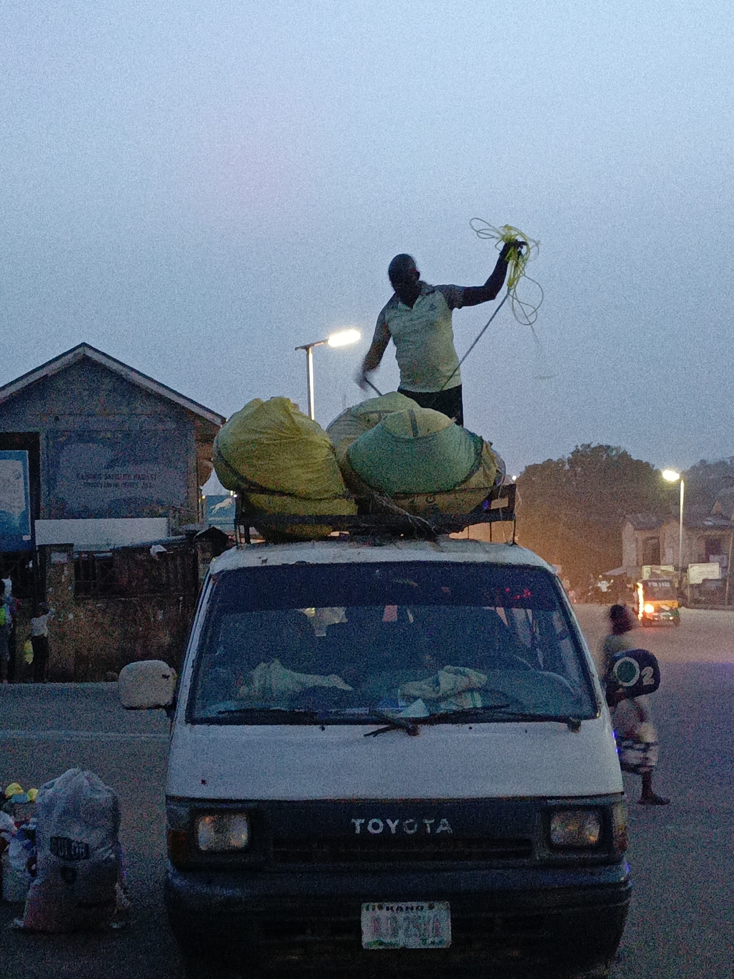 A medium shot captures a dusty white Toyota van heavily laden with large sacks secured to its roof. A man stands atop the vehicle, holding yellow rope, presumably to tie down the load. The license plate reads 'DEKANO 9', identifying the van's origin. The lighting suggests it's either dawn or dusk, as the sky is overcast. To the left of the van is a makeshift building, and several bags and other items are at the front corner of the van. To the right, another person is standing on the side of the road. In the background, there's another van and buildings lined by streetlights. The environment has an urban feel, suggesting a market or roadside commerce area. The location is the city of Unknown, Unknown.