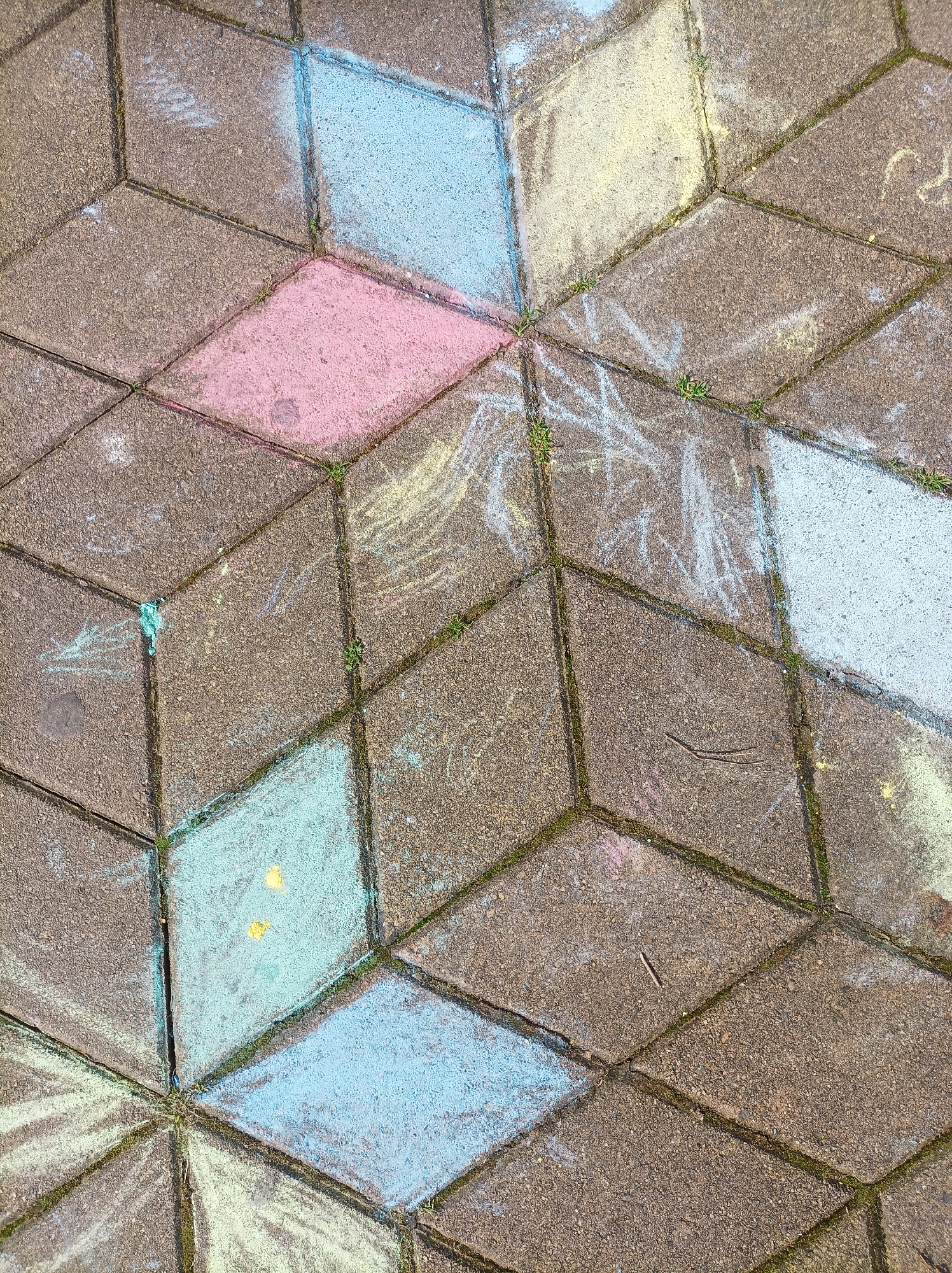 The image is a close-up shot of a paved walkway or patio. The paving stones are arranged in a diamond or rhombus pattern, creating a three-dimensional effect. Some of the stones are colored with chalk in shades of pink, blue, yellow, and white. Scrawls of chalk are also seen on some of the uncolored stones. Small bits of vegetation are growing in the cracks between the stones. A small twig is lying on one of the plain paving stones. The setting is possibly in Batumi, Georgia, as requested, but not definitively indicated in the image itself.