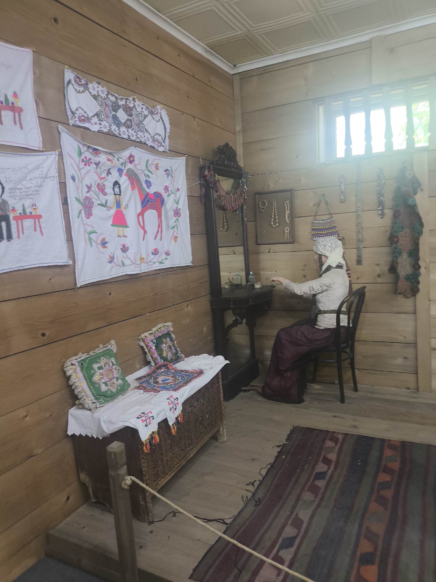 This image shows a room in the Ethnographic Museum of Adjara in Batumi, Georgia, designed to resemble a traditional Svan house interior. The walls are made of light-colored wood planks. On the left wall, several embroidered cloths are displayed, one featuring a stylized person and a red horse. In the foreground, a wooden chest is covered with white fabric, topped with two decorative pillows and another embroidered cloth. A rope barrier separates the viewer from the exhibit.
In the background, a mannequin dressed in traditional clothing, including a white headscarf, sits at a dark wooden vanity table with a mirror. The mannequin is reaching towards the table. Various decorative items, including necklaces and a framed display of wooden combs and pendants, are hung on the wall behind the mannequin. A window with vertical wooden slats is visible on the right wall, allowing bright light to enter. A patterned rug with stripes and geometric designs lies on the wooden floor in the foreground. The ceiling is covered with textured tiles.