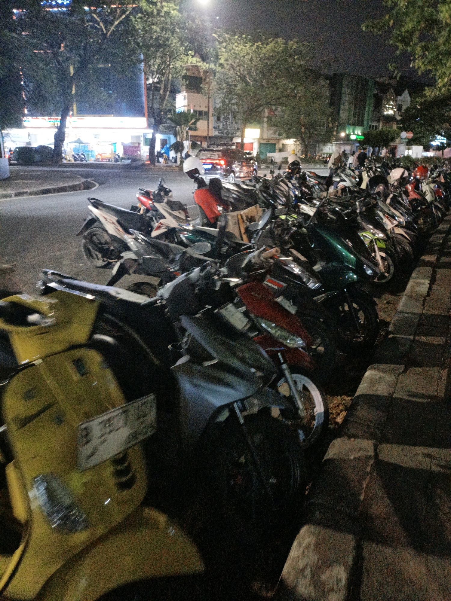 The image depicts a street scene at night in West Jakarta, Indonesia. A prominent feature is a long row of parked motorcycles lined up along a sidewalk. The motorcycles vary in color and style, with many appearing to be scooters. In the foreground, a bright yellow scooter with a license plate is visible. In the background, buildings with illuminated windows and storefronts suggest an urban environment. Trees with dense foliage are also present, and streetlights cast a glow on the scene. The overall impression is of a busy area with a lot of motorcycle traffic, likely a common sight in Jakarta, especially in the evening. There are no visible people actively interacting or engaged in specific actions, but their presence is implied by the parked vehicles. The image is characterized by low light conditions, typical of a nighttime urban setting.