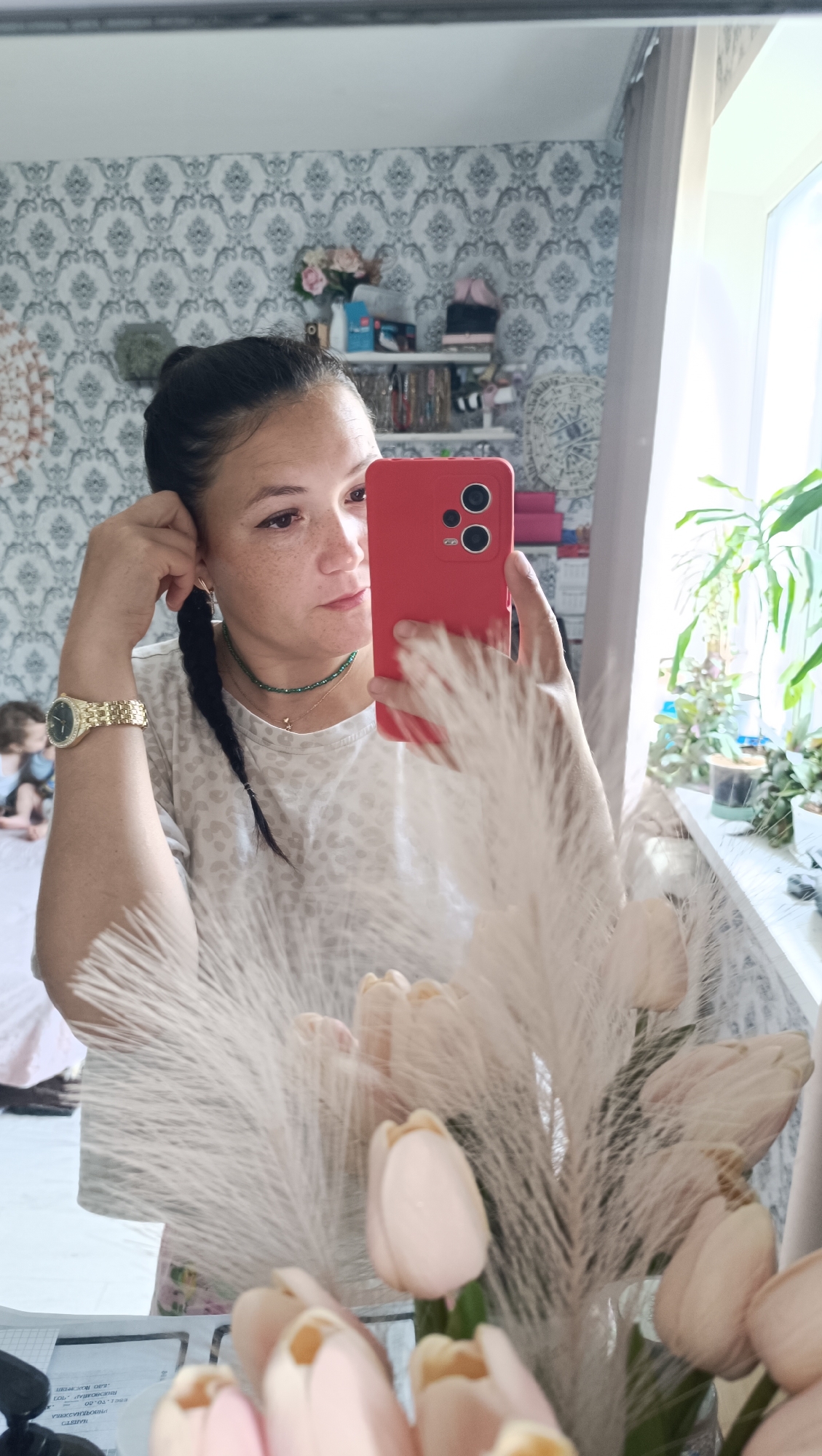 A woman with dark hair in a braid is taking a mirror selfie in a room. She is wearing a white t-shirt with a leopard print pattern, a green necklace, and a gold watch. She is holding a red smartphone in her right hand. In the background, there is a shelf with various items, including flowers, and a window with plants. There is also a child in the background. The room has patterned wallpaper.
