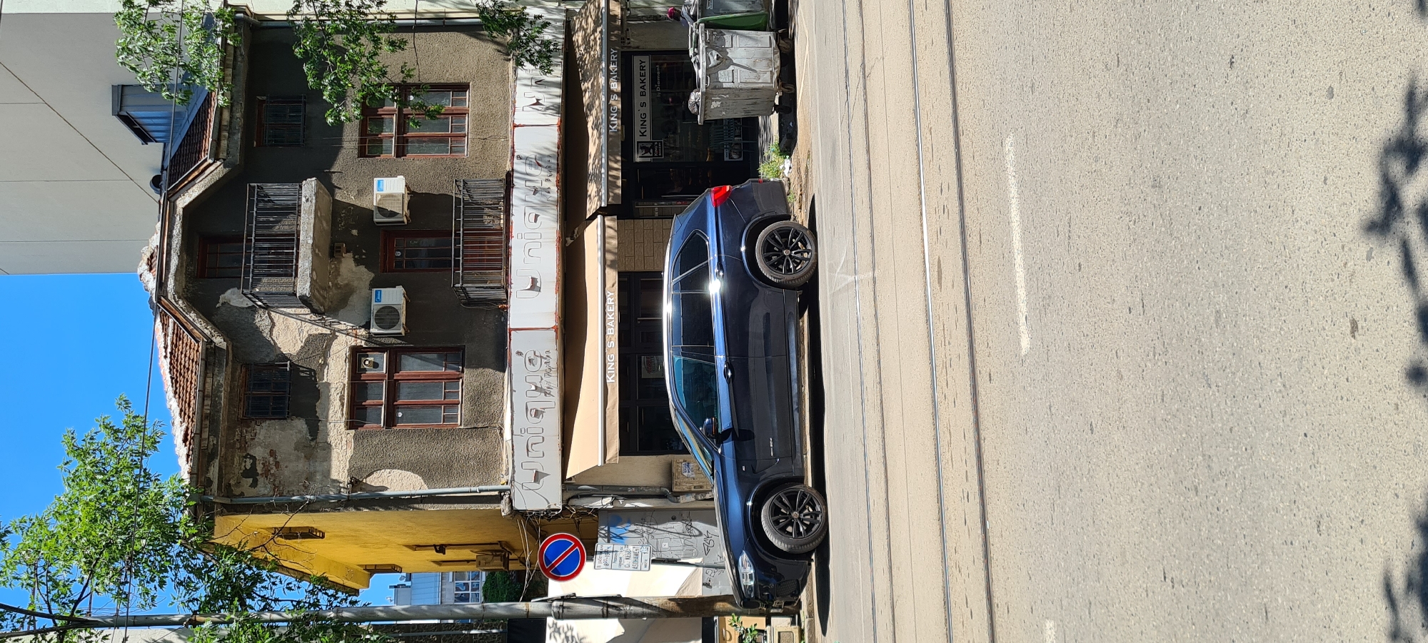 A dark blue BMW X5 SUV is parked on a street in Sofia, Bulgaria, parallel to a multi-story building. The building appears to be old, with weathered paint and a damaged roof. A bakery called "King's Bakery" is located on the ground floor of the building, with awnings extending over the entrance. In front of the building, there are several large, metal trash cans. The street is paved and has tram tracks. The sky is clear and blue, and trees with green leaves are visible on the left side of the image. A blue and red circular traffic sign is also visible on the left side of the image.