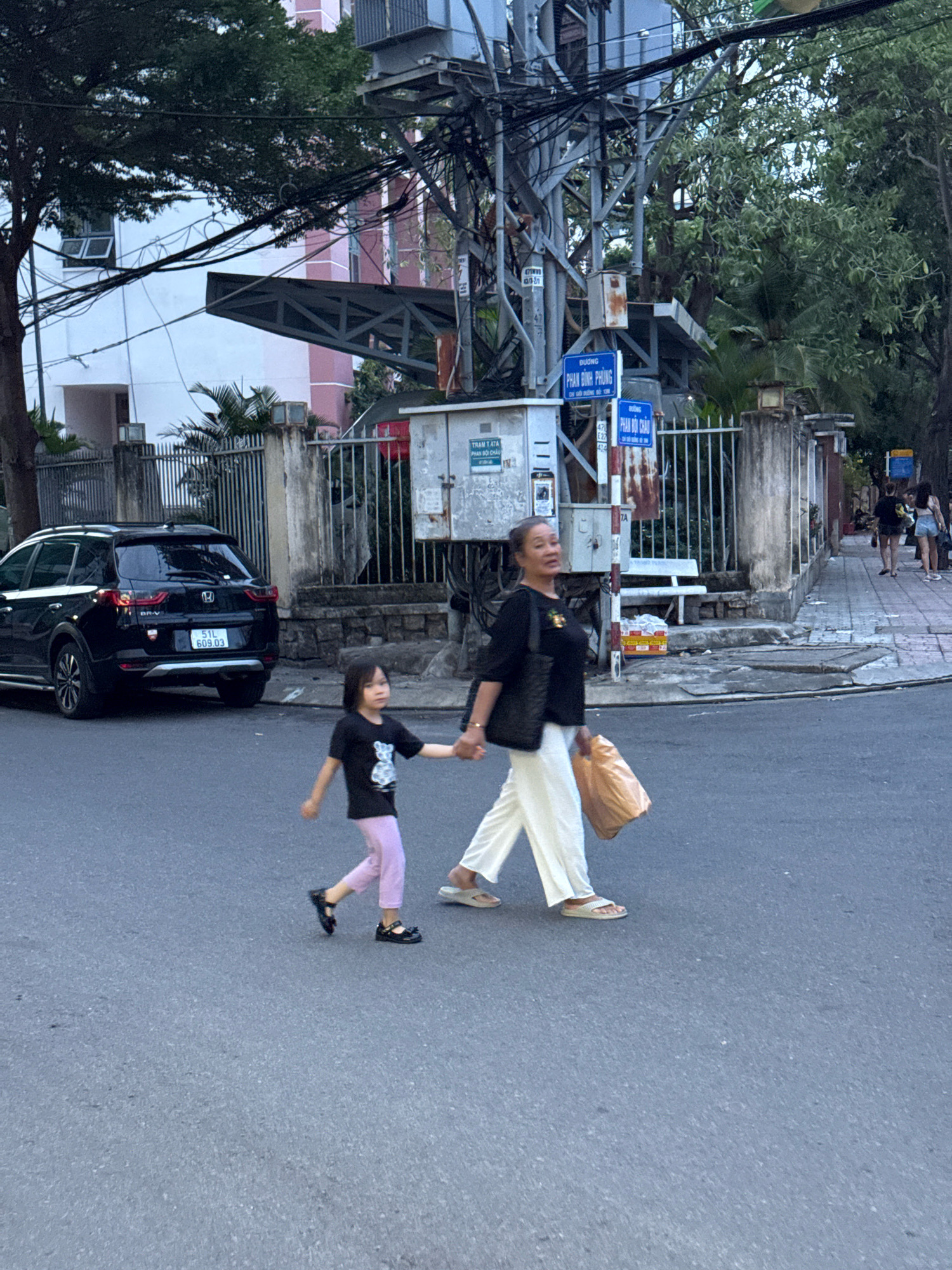 An adult woman and a young girl are walking across a paved street in Nha Trang, Vietnam, holding hands. The woman, on the right, is wearing a black shirt, white pants, and sandals, and is carrying a large brown bag. The girl, on the left, is wearing a black t-shirt and light pink pants, and black shoes. Behind them, a black SUV is parked on the side of the road. In the background, a utility pole with numerous wires is visible, along with a fence and buildings. Signs with Vietnamese text are attached to the utility pole.