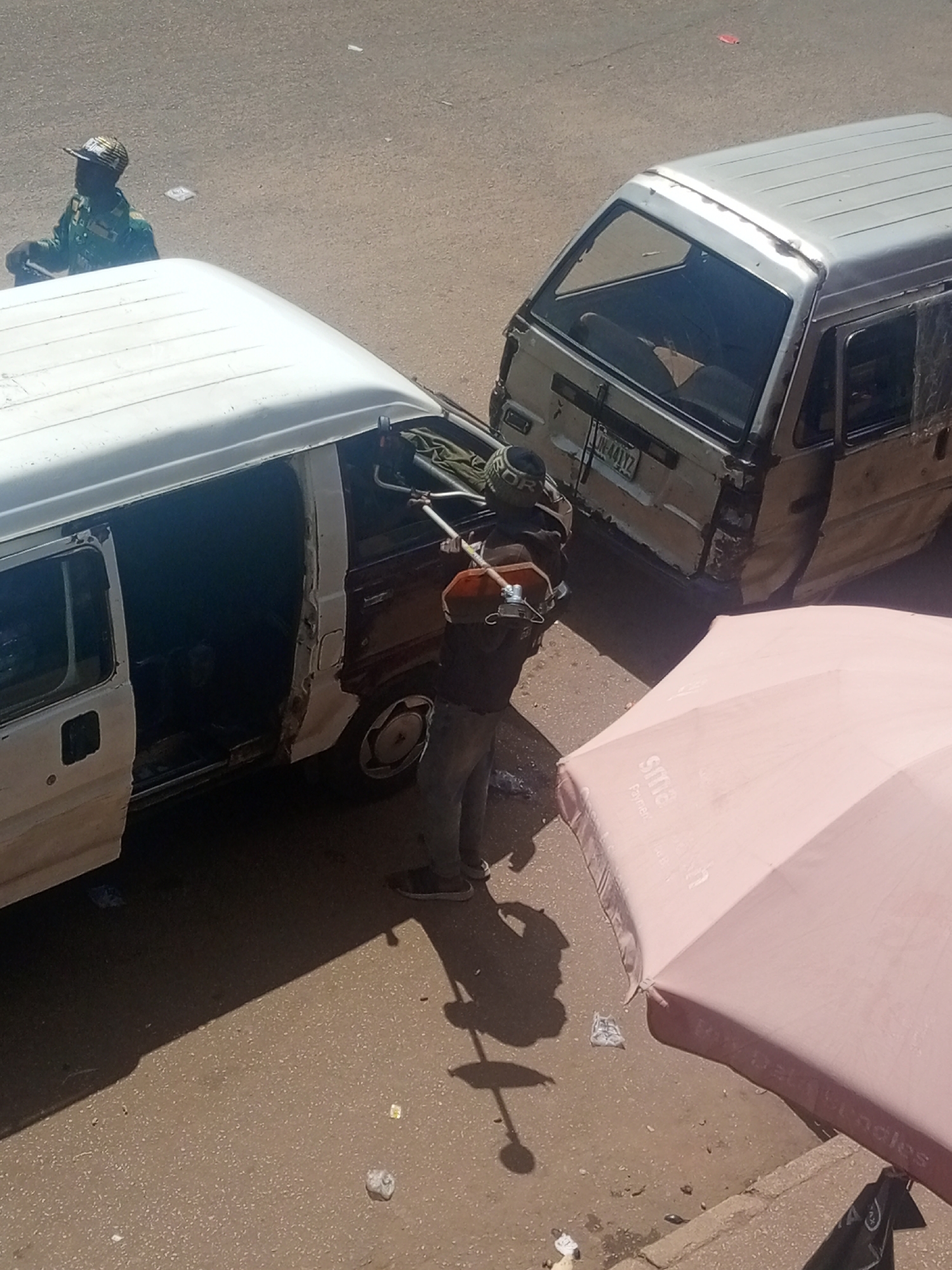The image shows two vans parked on a dusty, paved surface, likely a roadside. The van in the foreground is white with its doors and panels removed, appearing to be undergoing repairs or being dismantled. A person, dressed in dark clothing and a cap, is standing next to it, holding what looks like a tool or part of the vehicle. Another person in a green uniform and a patterned cap is visible in the background, standing near a motorcycle. The second van, a lighter color, is parked behind the first and also shows signs of damage or wear. A large, pink umbrella is partially visible in the lower right of the frame, casting a shadow on the ground, suggesting it's daytime and sunny. The ground is scattered with debris, typical of an outdoor environment. The license plate on the second van is partially visible, reading "JN-441YZ". The overall scene suggests a busy, possibly informal, repair or dismantling operation in an outdoor setting. The location is identified as Bukuru, Nigeria.