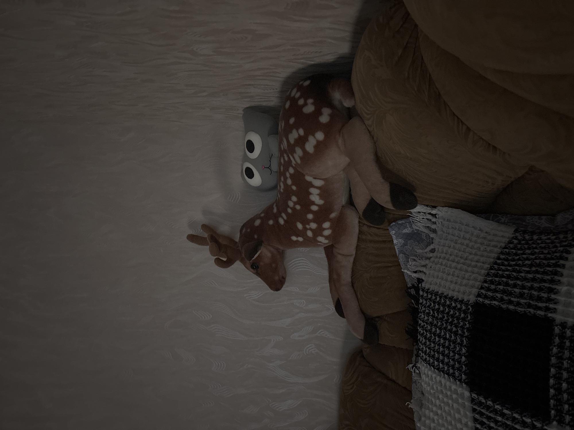 The media file contains a stuffed deer and a stuffed toy with large eyes, both lying on a surface. The stuffed deer is brown with white spots and has antlers. The other stuffed toy is gray with two large black eyes and a small mouth. They are positioned next to a textured, light-colored surface that appears to be a carpet or fabric. To the right of the stuffed animals is a tan-colored upholstered object, possibly a chair or couch, with a textured pattern. A blanket with a black and white checkered pattern is draped over the tan upholstery. The lighting in the scene is dim, suggesting it might be nighttime or a poorly lit room. The overall impression is a still life arrangement of toys. There is no discernible activity or event taking place. No visible text is present in the media file. The provided location context of Sinyavino, Russia, does not offer any visual cues within the image itself to confirm or deny this setting.