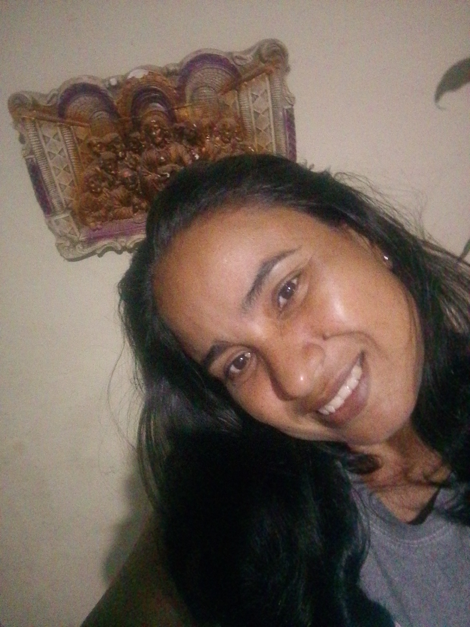 A selfie of a person with long dark hair smiling. The person is wearing a grey t-shirt. Behind the person is a decorative, framed art piece hanging on the wall. The image was likely taken indoors. There is no indication of the location in the city of Juan Griego, Venezuela.