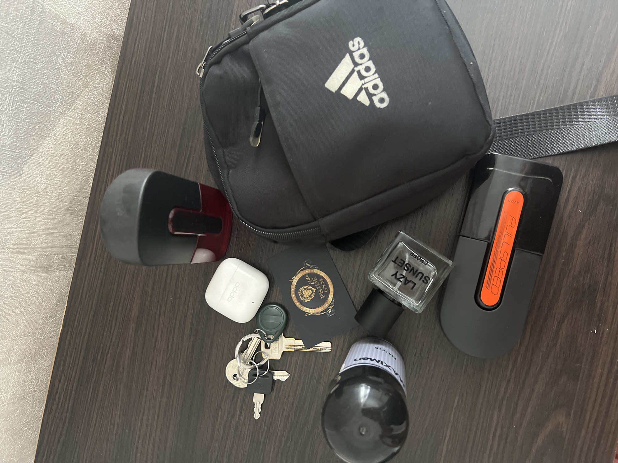 The image displays a collection of personal items laid out on a dark wooden surface, likely a desk or table. The items are arranged in a somewhat scattered manner. In the foreground, there is a black cylindrical object with a white label, a glass perfume bottle with a black cap and the label "LAZY SUNSET," and a black electronic device with an orange accent. Behind these, a set of keys with various keychains is visible, alongside a white wireless earbud charging case. To the right, a black crossbody bag with the Adidas logo is prominent. In the upper portion of the image, a black and red object, possibly a stamp or a dispenser, is positioned. The background appears to be a textured wall. The scene is static, with no discernible actions or interactions taking place. The lighting suggests an indoor environment. The location is not visible in the image, and there is no information to suggest it is in Sobishchytsi, Ukraine.