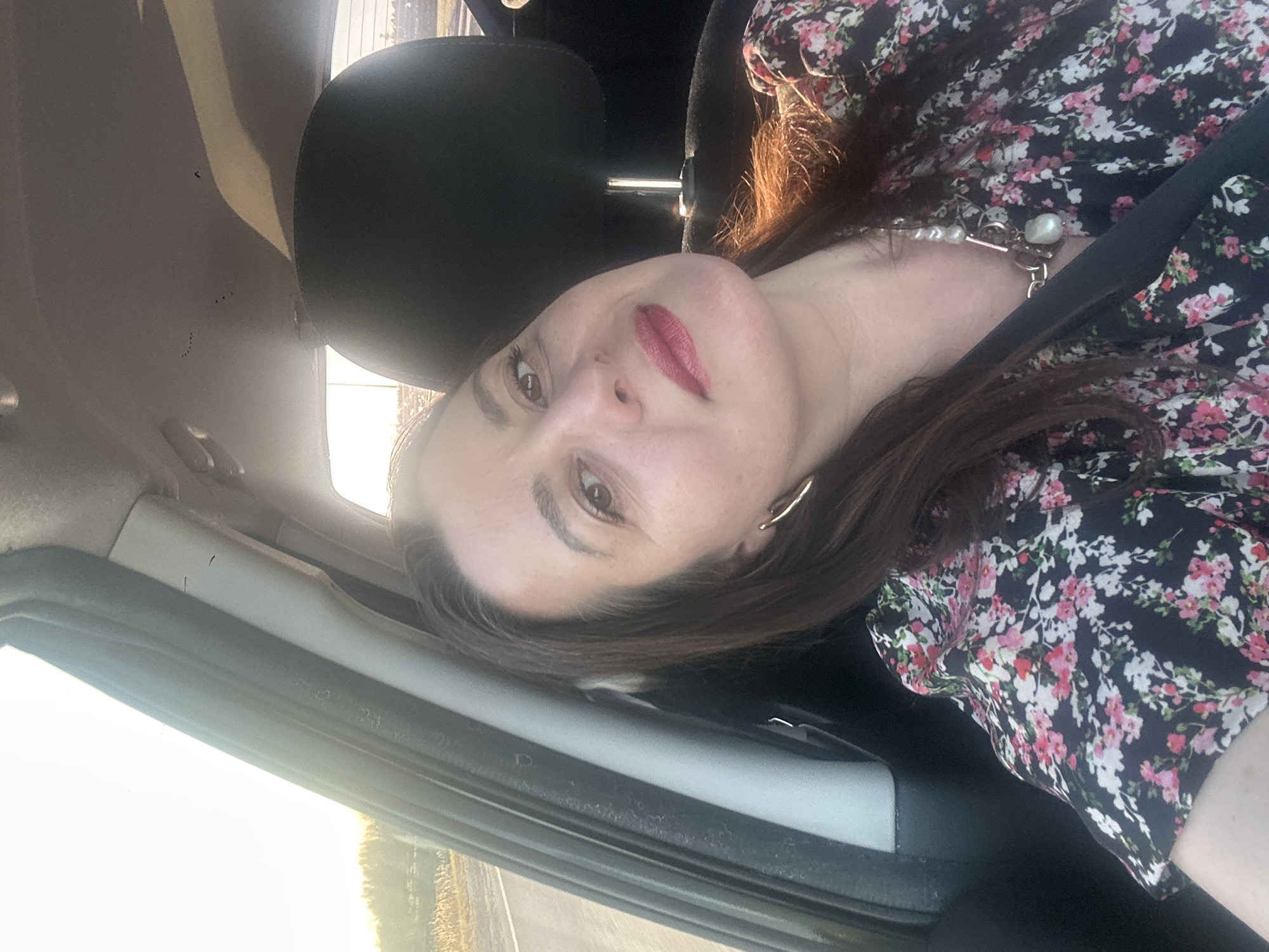 A woman with brown hair and light skin is sitting in the driver's seat of a car. She wears a floral-patterned blouse and a pearl necklace. The car interior is visible, including the black headrest and the car's frame. Outside the window is a bright, hazy scene suggesting either sunrise or sunset. The location is specified as the city of Piņķi, Latvia, but this is not visually verifiable from the image.
