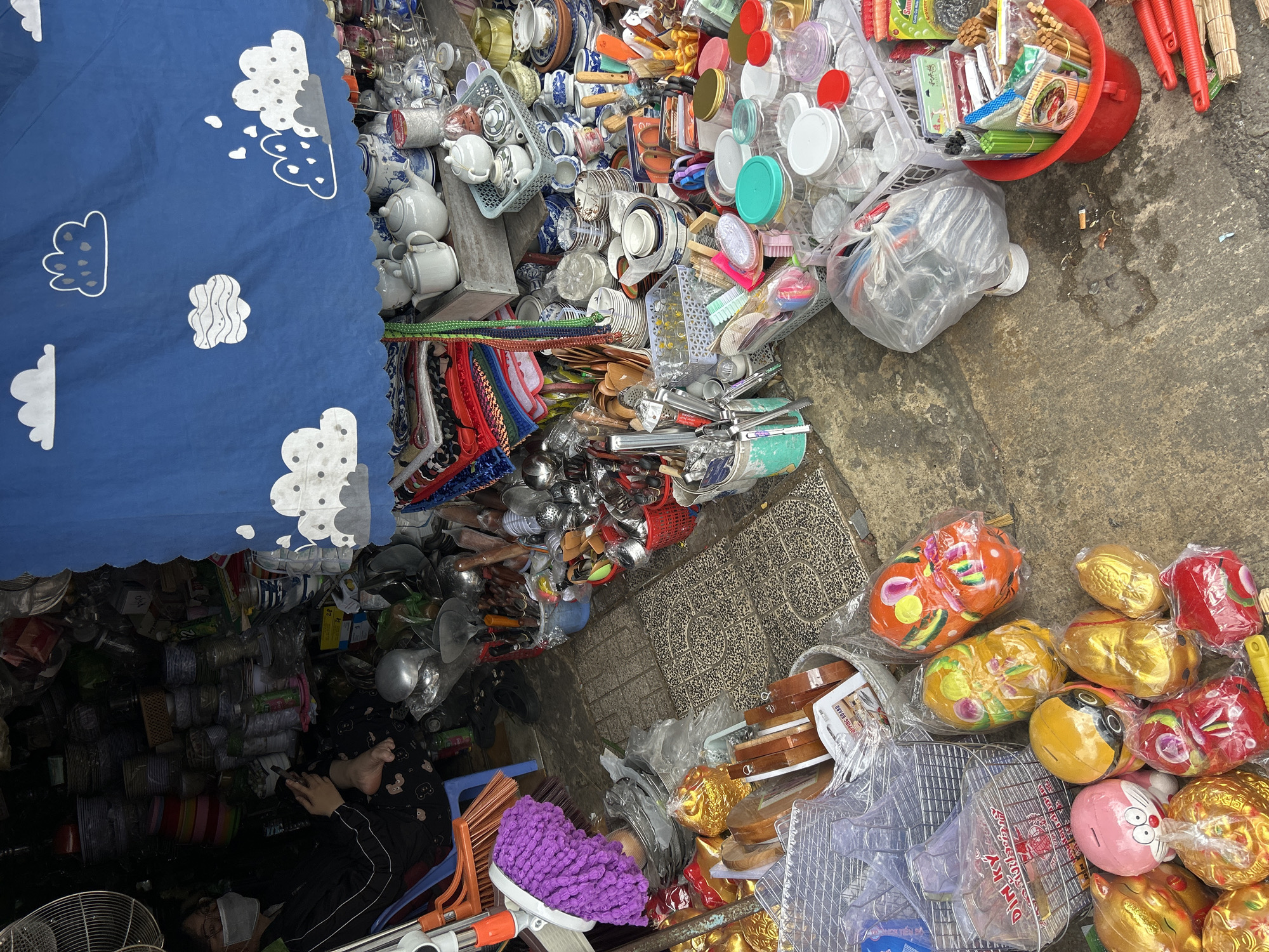 This overhead shot captures a densely packed outdoor market stall in Nha Trang Ward, Vietnam, showcasing a wide array of household goods. The scene is bright, suggesting daytime, though a blue tarp with white cloud patterns provides shade over part of the stall.
The primary subject is a market vendor, visible on the left side, sitting barefoot on a blue plastic chair. They wear dark clothing, possibly patterned pants, and a face mask, appearing to be engrossed with a mobile phone.
The stall is overflowing with countless objects. On the upper left, there's a large collection of ceramic items, including white teapots, cups, and blue-and-white patterned dishes, some neatly stacked in plastic baskets. Adjacent to these are numerous plastic containers with colorful lids, brushes, and various kitchen utensils like spatulas and scoops. Further down, bundled items like colorful ropes or straps are visible.
The ground area displays more goods and details. A red plastic bucket holds chopsticks and skewers. Scattered on the concrete pavement are discarded cigarette butts, hinting at a public space. Towards the bottom right, a collection of novelty piggy banks, some in gold and others brightly colored animal shapes (like cats, fish, and pigs), are individually wrapped in plastic. Metal racks and wooden cutting boards are also part of the merchandise. Cleaning supplies, such as a purple microfiber mop head, are also present. The ground is a mix of concrete and patterned tiles.
Visible text includes "DINH KY" on a clear plastic package or rack, "SUNNY..." on a green product wrapper, and "OVER LOAD" on a brown cardboard package. The overall impression is a bustling, yet momentarily quiet, market environment filled with practical and decorative items for daily life.