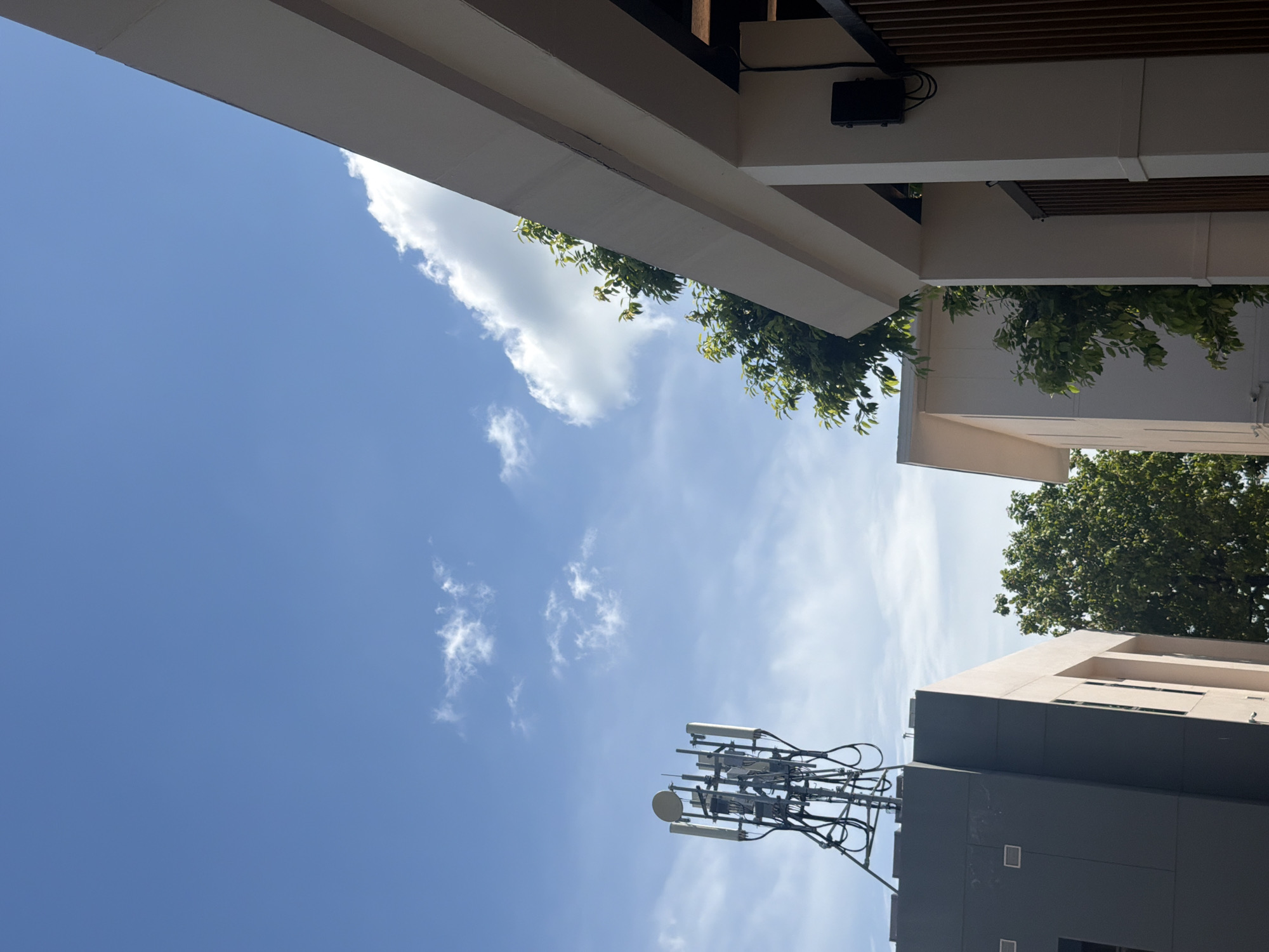 The image shows a communication tower on top of a building in Baan Chaweng, Thailand. The sky is blue with some clouds. There are trees and other buildings in the background. The communication tower has several antennas. The building in the foreground is white with a beige trim.