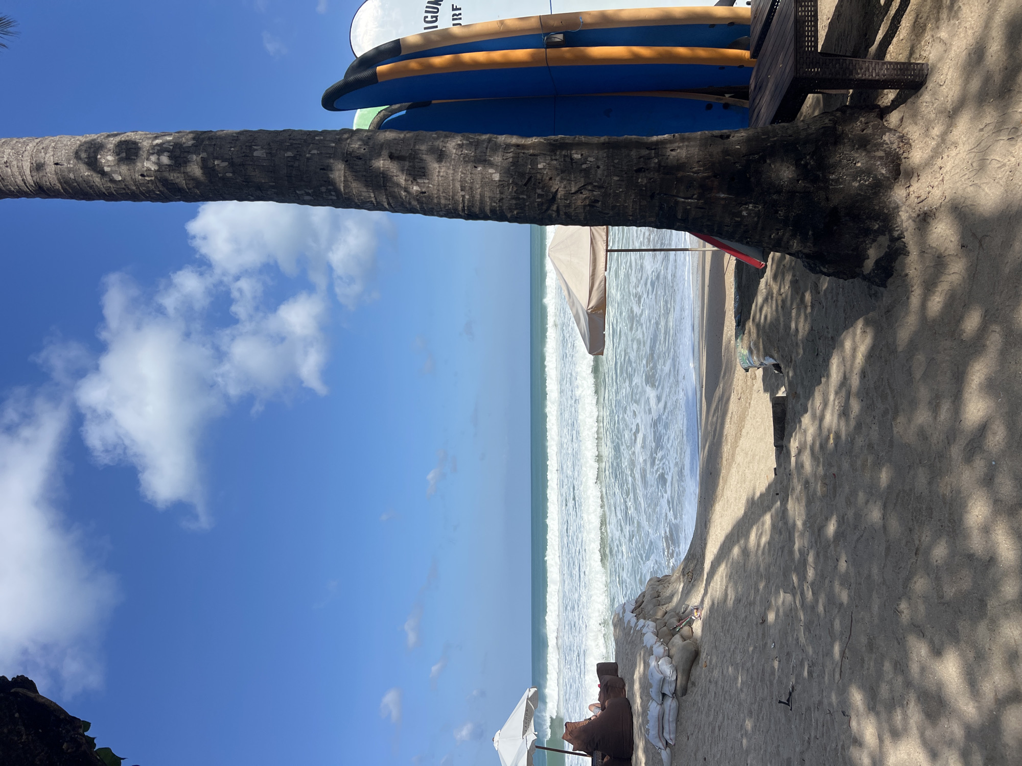 This image depicts a sunny day at a beach in Kuta, Indonesia. The foreground is dominated by a sandy beach with the dappled shadows of palm fronds cast across it. A large palm tree trunk stands prominently on the right side of the frame, with several surfboards leaning against it. These surfboards are varied in color, including blue, orange, and white. A dark brown table is placed next to the palm tree. Further into the scene, a beige umbrella shades a seating area on the sand. Two brown beanbag chairs are positioned beneath a white umbrella, where a person is seen relaxing, facing the ocean. The ocean itself is a vibrant blue, with gentle waves rolling towards the shore. Sandbags are stacked along the edge of a slightly elevated sandy bank that separates the beach seating area from the water. The sky above is a clear blue with scattered white clouds. The overall atmosphere is one of a tranquil beachside resort.