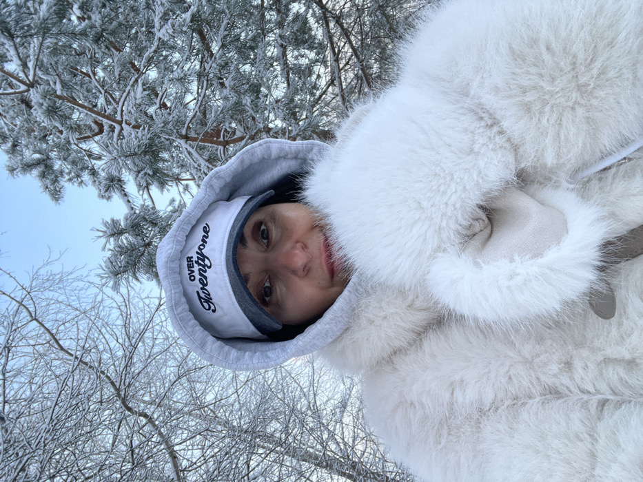 A female subject is captured in a close-up selfie outdoors in a snowy, wooded area of Syasstroy, Russia. She looks directly at the camera with a neutral expression, her cheeks appearing flushed. She wears a white baseball cap with a blue brim, featuring "OVER TwentyOne" embroidered in black script, covered by the pulled-up hood of a grey sweatshirt. A thick, white faux fur coat covers her upper body. Her left hand is partially visible, clad in a light-colored mitten, appearing to hold the fur of her coat. Her lips are covered with reddish-pink lipstick. The background consists of deciduous trees with snow-dusted bare branches and coniferous trees heavily laden with snow or frost, all beneath a pale blue sky.