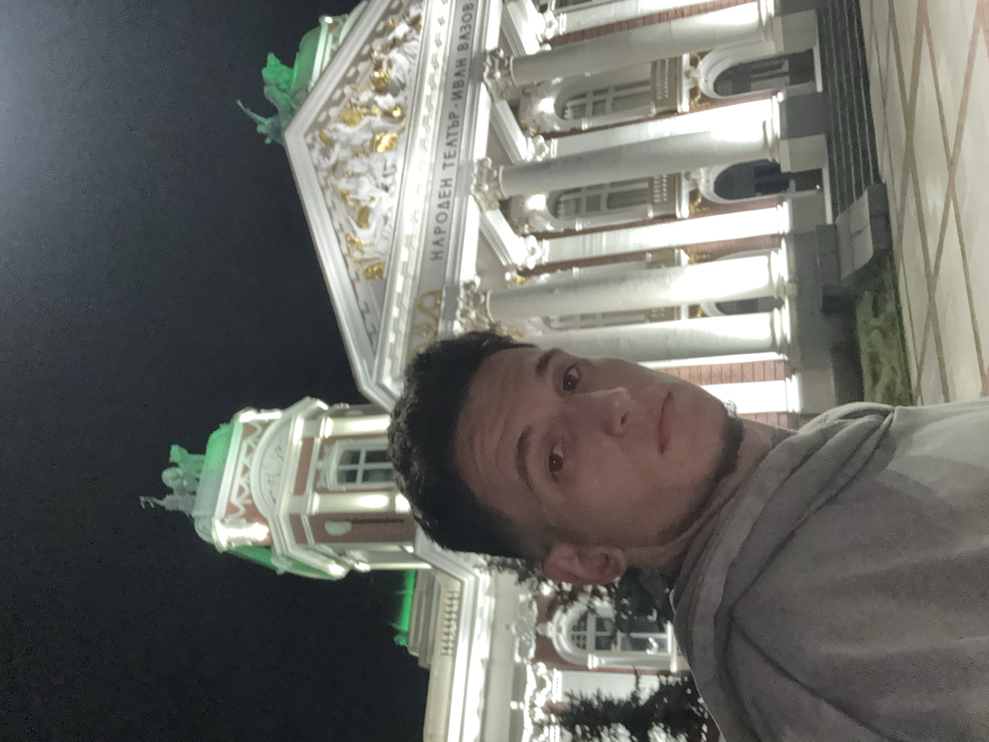 A person is taking a selfie at night in front of the Ivan Vazov National Theatre in Sofia, Bulgaria. The building is illuminated, showing its architectural details, including columns, inscriptions in Bulgarian, and a sculpture on the roof. The sky is dark. The person is wearing a gray shirt.