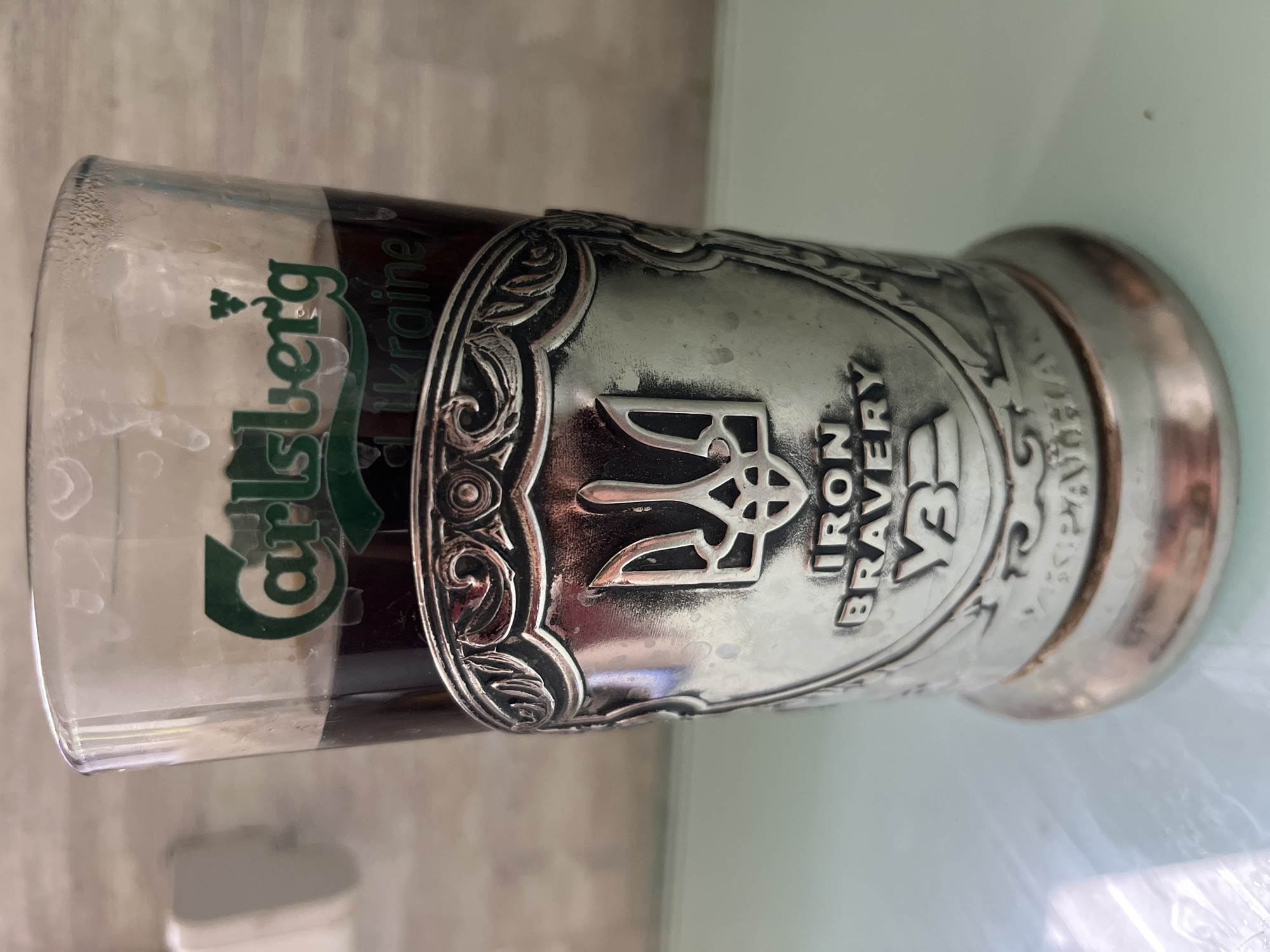 The image shows a close-up view of a glass of dark liquid, likely beer, adorned with a decorative metal holder. The glass has the "Carlsberg Ukraine" logo printed in green on its upper section. The metal holder is intricately designed with ornate patterns and features a prominent symbol resembling a trident, the national emblem of Ukraine, in its center. Below the trident, the words "IRON BRAVERY" are embossed, along with the Cyrillic letters "УБ". The holder is attached to the base of the glass, which appears to be made of a metallic material. The overall scene is well-lit, with the background appearing to be a textured wall. The image does not provide any information about the specific location within Kharkiv, Ukraine.
