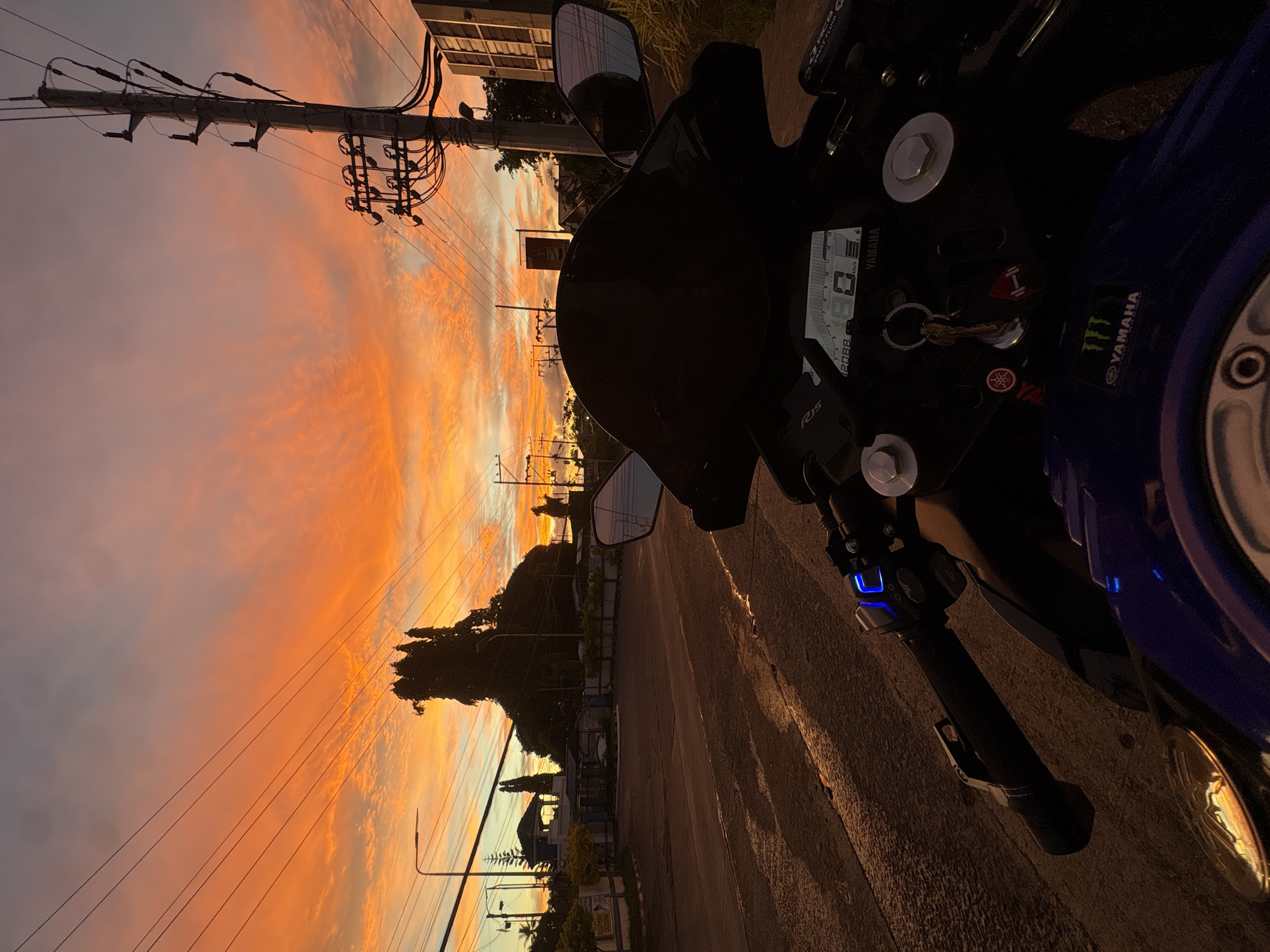 This is a low-angle, first-person perspective photo taken from a motorcycle, likely in Davao City, Philippines, during a vibrant sunset. The sky is ablaze with fiery orange and red hues, accented by streaks of pink and purple clouds. Power lines and utility poles crisscross the sky, silhouetted against the dramatic backdrop.
In the foreground, the motorcycle's handlebars and instrument cluster are visible. The digital display shows speed and other indicators. A set of keys is hanging from the ignition. The motorcycle's dark bodywork contrasts with the bright sky.
Beyond the motorcycle, a road stretches into the distance, wet and reflective, hinting at recent rain. The road is lined with trees and some buildings. A notable structure with a blue roof and white walls is visible on the left side of the road. Streetlights are also present. The overall atmosphere is serene, with the fading daylight creating long shadows. No people are visible in the frame.