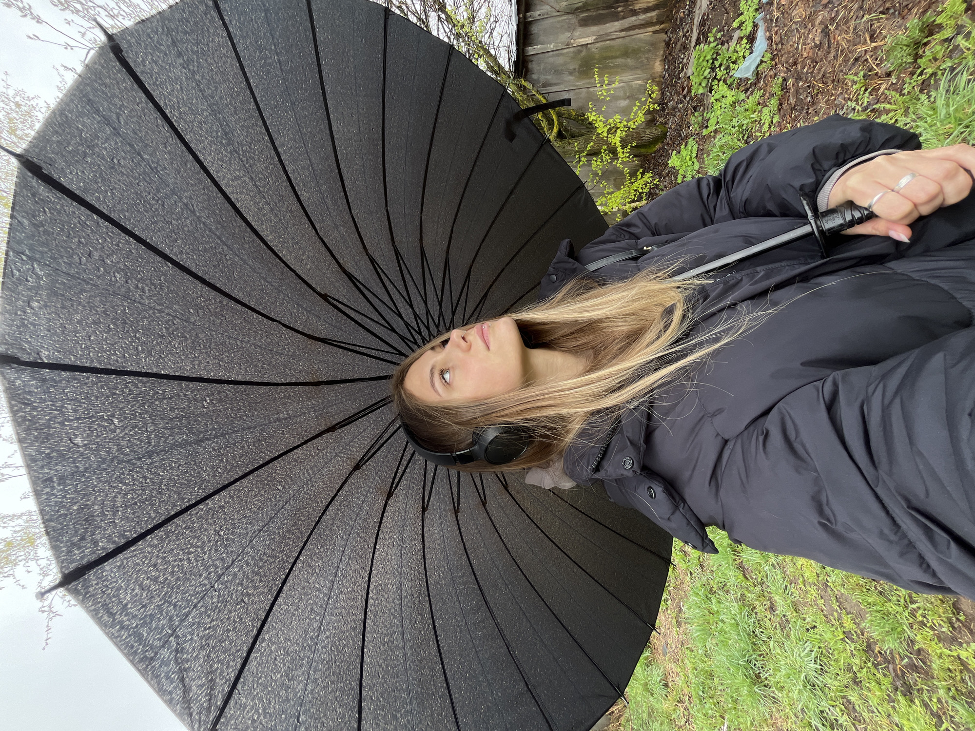 A young woman stands outdoors, holding a large black umbrella. She wears a black puffer jacket and headphones. The background includes green grass, a wooden fence, and some foliage. The image is taken from a low angle, looking up at the umbrella and the woman's face. Фоки, Russia cannot be confirmed based on the visual information provided.