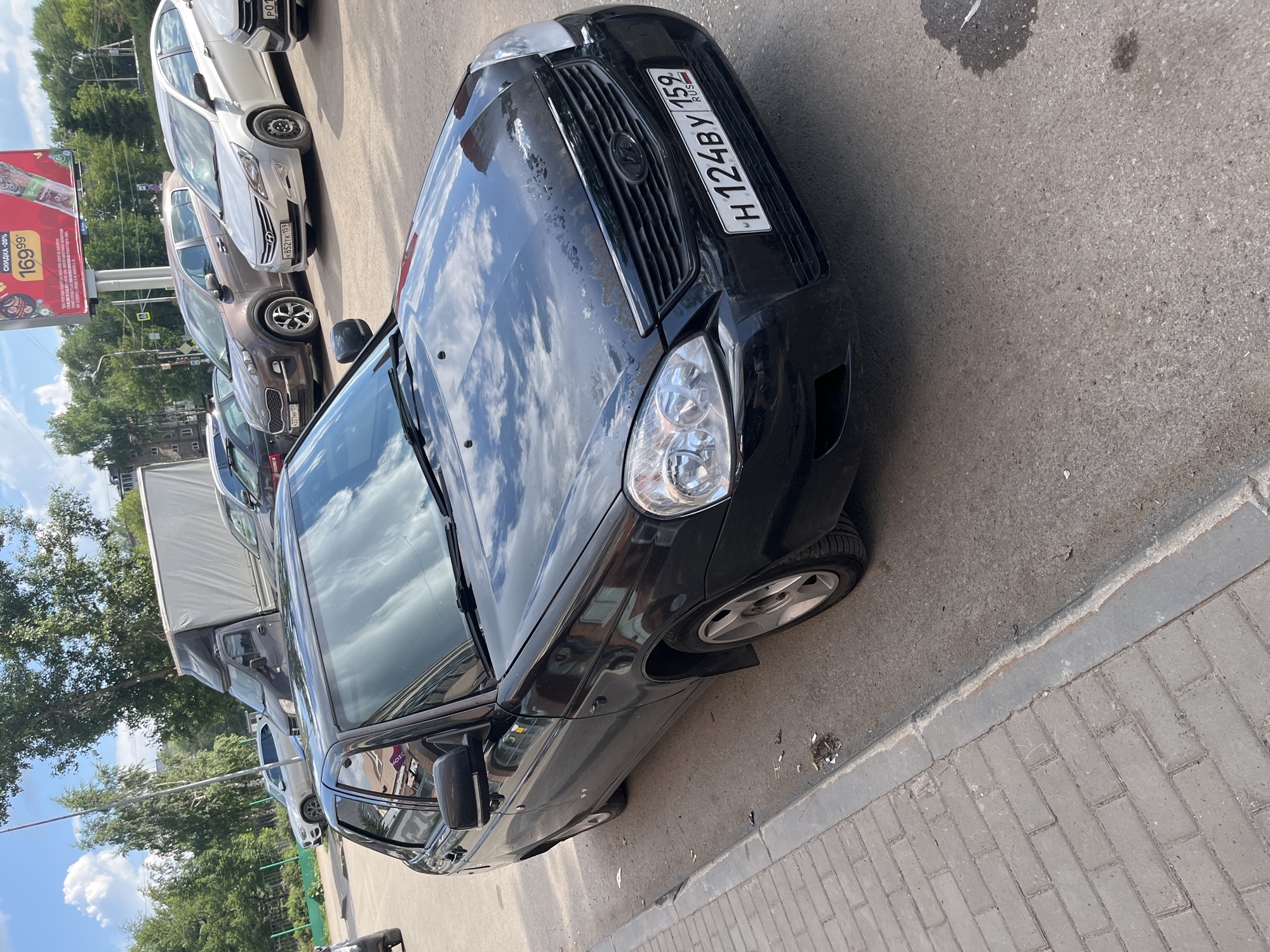 A black sedan is parked on a paved street adjacent to a sidewalk in Perm, Russia. The car is positioned at an angle, with its front passenger side facing the viewer. The license plate reads "H 124BY 159". Several other vehicles are visible in the background, including a white SUV, a brown hatchback, and a white sedan. A large billboard advertising a sale is also present in the background. The sky is blue with scattered clouds, and trees line the street. The scene appears to be a typical urban environment in Russia.