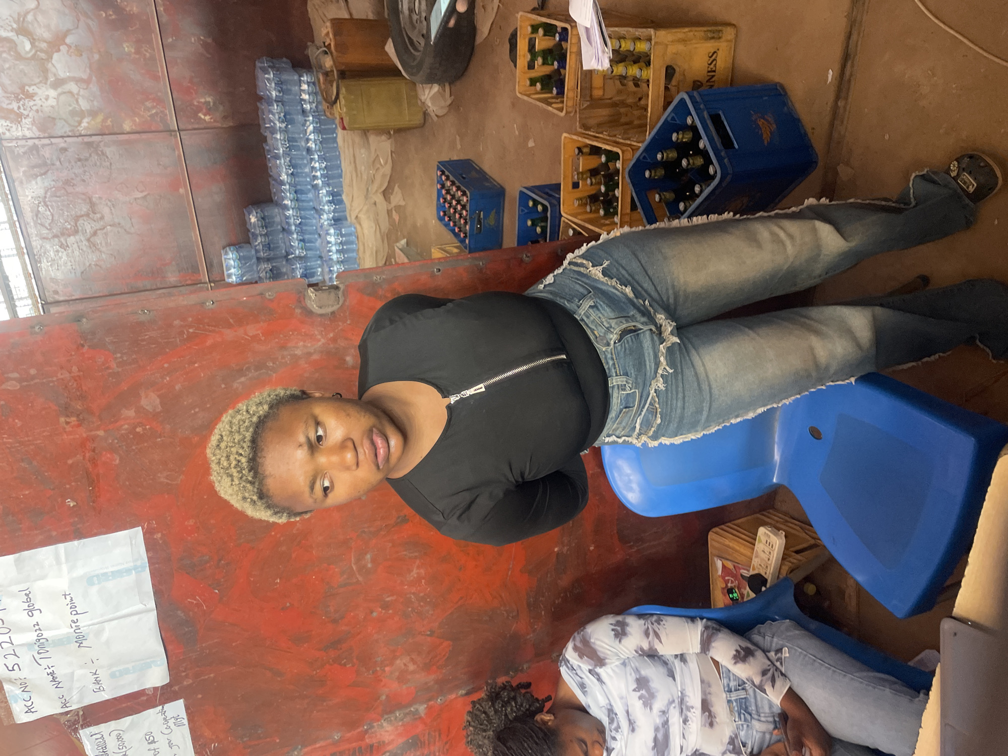 The image shows two individuals inside a container or truck in Gura Top, Nigeria. One person with short, dyed-blonde hair is sitting upright on a blue plastic chair, facing the camera. This person wears a black zip-up shirt and light-wash denim jeans. Another person with dark, curly hair is reclined on another blue plastic chair. They are wearing a light-colored, patterned top and jeans. Several bottle crates, some full of dark bottles, are scattered on the floor around them. There are also stacks of water bottles. In the upper left corner, a piece of paper with handwritten text is partially visible, with the text "ACC NO: 52205" and "Acc NAME: Tonigozz global". The interior walls appear to be made of metal.