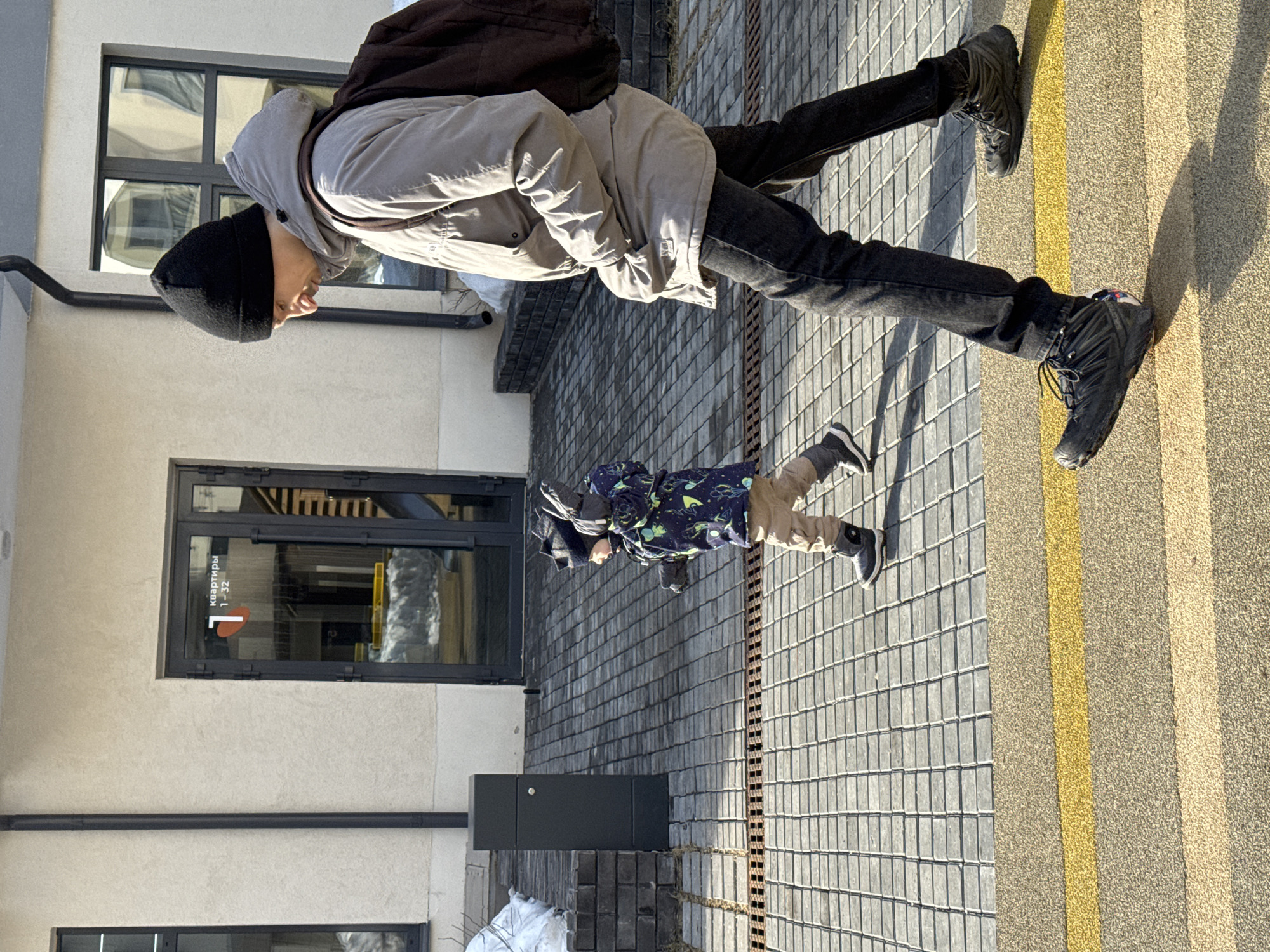 The image displays an adult male and a young child walking leftward on a paved pedestrian area adjacent to a building in Izhevsk, Russia. The scene is illuminated by bright daylight, casting distinct shadows to the right, with patches of dirty snow visible on the ground near the building, indicating cold weather conditions.
The adult male, positioned to the right of the frame, wears a black knit beanie, a light grey hooded winter jacket over a brown backpack, dark jeans, and dark athletic shoes. His head is slightly bowed, and his right hand is in his jacket pocket.
The young child, positioned slightly ahead and to the left of the adult, wears a dark winter jacket with a multi-colored pattern, light brown pants, dark winter boots, a dark knit hat with ear flaps, and dark gloves. Both of the child's arms are bent at the elbows and slightly raised.
The ground consists of grey square pavers, with a section in the foreground featuring a textured light brown surface crossed by a bright yellow horizontal stripe. A metal drainage grate is embedded in the pavement. The background features a light-colored stucco building with multiple dark-framed windows and a dark-framed glass entrance door. The entrance door displays a red stylized "T" logo and the Russian text "квартиры 1 - 32," indicating apartments 1 through 32. A black vertical downspout runs down the building's exterior. To the left of the entrance, a dark grey rectangular pedestal or planter box rests on a low dark brick wall.