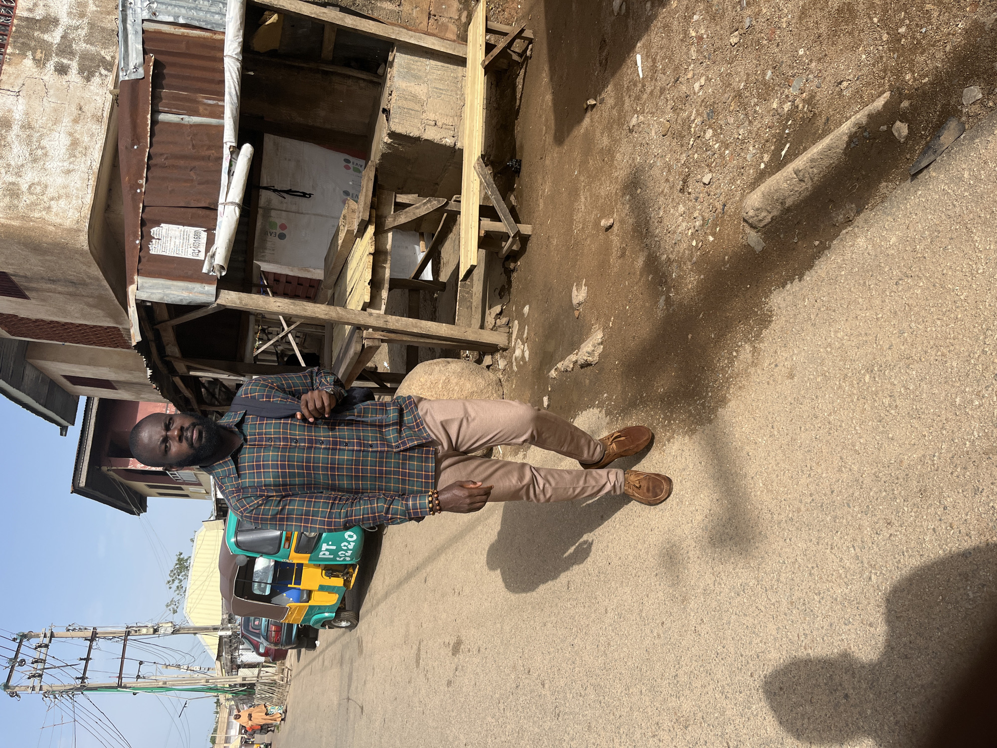 A Black man walks towards the camera on a paved road in Jos, Nigeria. He is wearing a plaid long-sleeved shirt, tan trousers, and brown shoes. He has a backpack on his right shoulder. To his left, a yellow and green motorized tricycle is parked. In the background, there are buildings and market stalls. The man is looking directly at the camera with a neutral expression. The scene appears to be a typical street in a busy urban area.