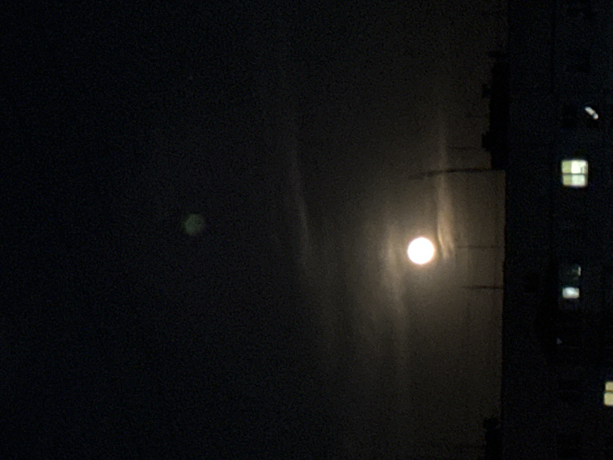 The image presents a dark, nighttime scene, likely an outdoor view. The primary subject is a bright, circular light source, strongly resembling the moon, positioned towards the right-center. This light emits a soft glow, illuminating the surrounding atmosphere with vertical streaks of light, suggesting the presence of haze or thin clouds. To the left of the moon, a faint, blurry, greenish-yellow oval shape is visible, which could be a distant light source or a lens artifact.
On the far right, a dark, angular structure, probably a building, partially occupies the frame. Within this structure, several rectangular windows are illuminated with a soft, yellowish light, contrasting with the prevailing darkness. The overall scene is characterized by very low light conditions, resulting in a grainy texture across the dark areas of the sky and background. There is no discernible activity taking place, and no visible text is present. The time of day is clearly night, with the illuminated windows hinting at human presence within the building.