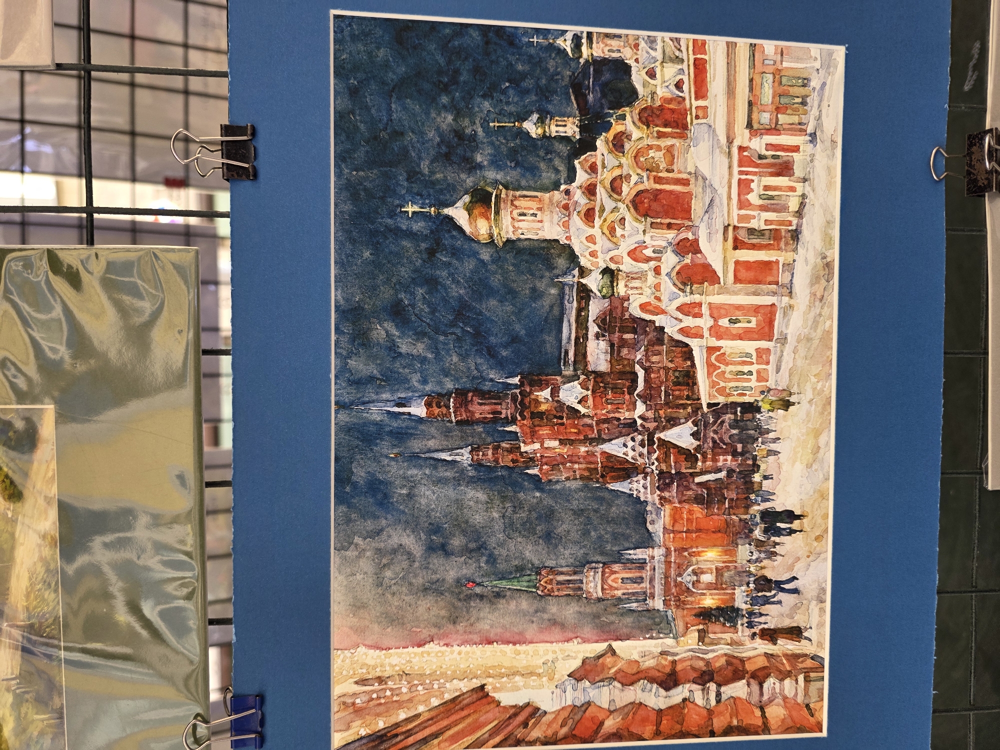 This media file is a watercolor painting of St. Basil's Cathedral in Moscow, Russia. The cathedral is depicted with its iconic colorful onion domes and intricate architectural details. The painting shows a busy scene with people walking and gathering in front of the cathedral. The sky is a deep blue, suggesting it might be late afternoon or early evening. The ground in front of the cathedral appears to be covered in snow, indicated by the white and light yellow hues. The overall mood of the painting is lively and vibrant, capturing the spirit of a bustling city square. There is no visible text in the image.