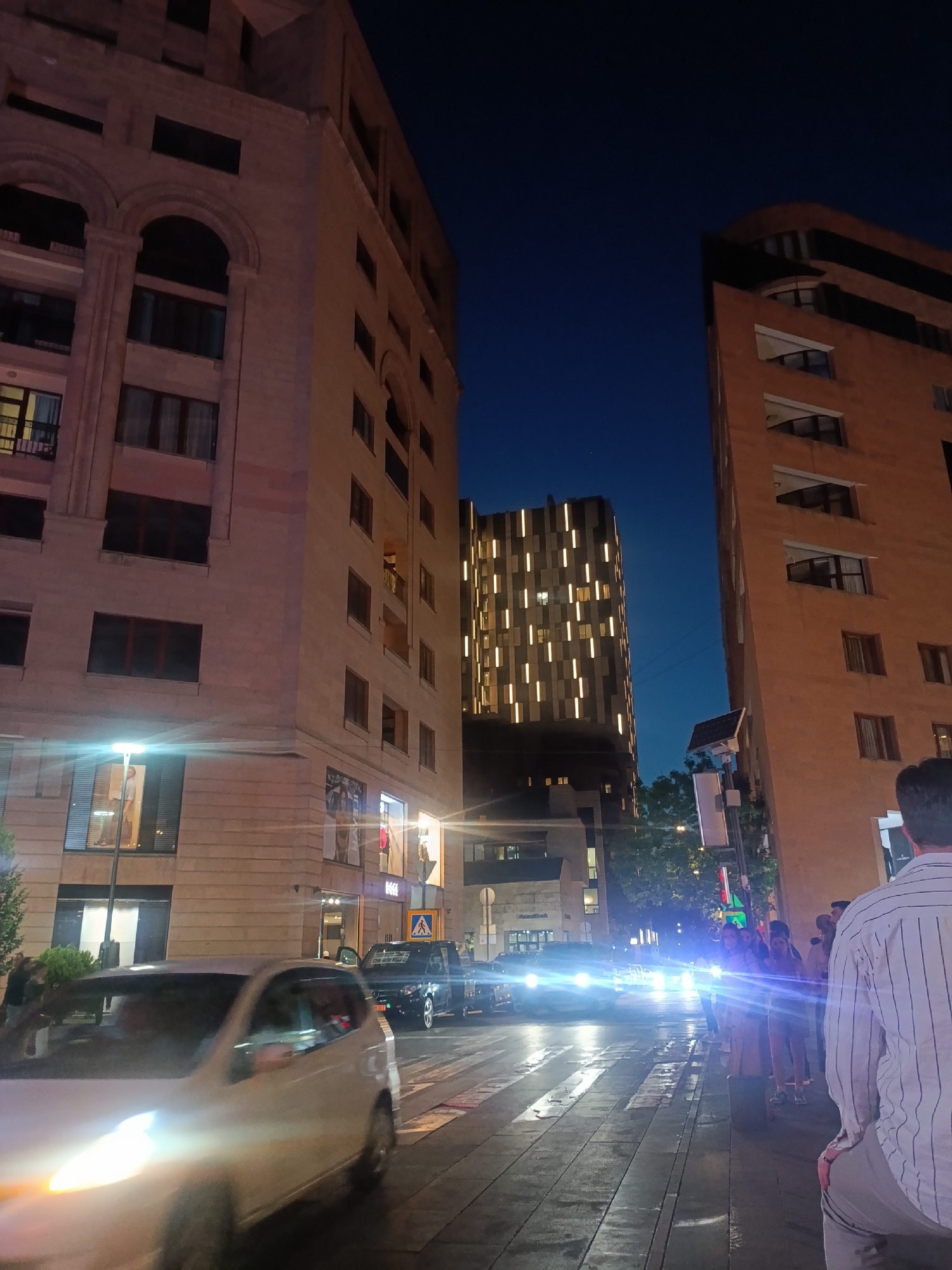 At night, a street scene in Yerevan, Armenia, shows buildings on either side of the street, with traffic and people on the sidewalks. On the left, a light-colored car drives toward the viewer, its headlights blurred due to motion. In the distance, car headlights create a bright glare. A group of people stands on the right sidewalk, some of whom are blurred. A person in a striped shirt stands in the foreground on the right side. A tall building with illuminated windows is visible in the distance. The sky is a deep blue. The street surface is wet, possibly from recent rain. Streetlights and building lights illuminate the scene.