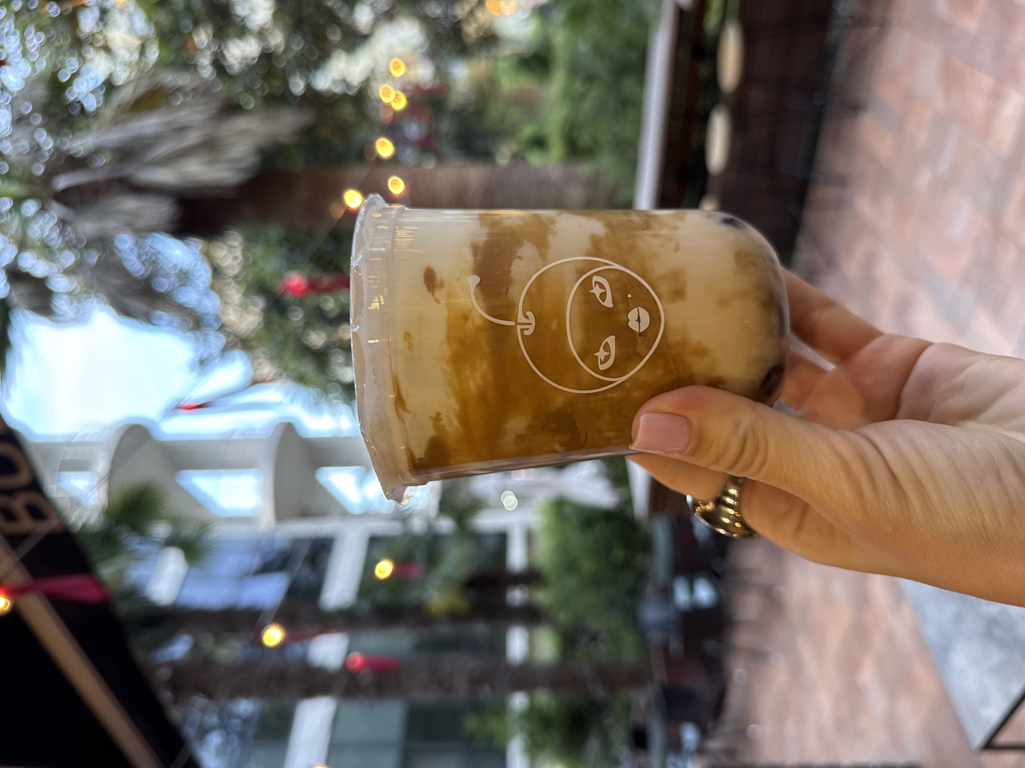 The image displays a close-up shot of a person's hand holding a clear plastic cup filled with a beverage, oriented horizontally across the frame. The drink inside is a creamy, light brown liquid with darker, marbled streaks, suggesting a layered concoction such as a bubble tea or a similar specialty drink. A distinctive white line-art logo is visible on the cup, featuring a stylized circular face with large eyes and a small mouth, connected to a curved line ending in an arrow that points towards the face.
The hand, with manicured nails painted in a soft pink, wears a golden ring on one finger. The background is softly out of focus, creating a vibrant bokeh effect. It depicts an outdoor setting with lush green foliage, including identifiable palm trees, and blurred outlines of building structures. Numerous warm, glowing string lights are scattered throughout the background, forming circular bokeh highlights, which suggest either a late afternoon or evening ambiance, or a well-lit shaded area. The scene evokes a casual and relaxed atmosphere, likely at an outdoor cafe or similar establishment in Sochi, Russia.