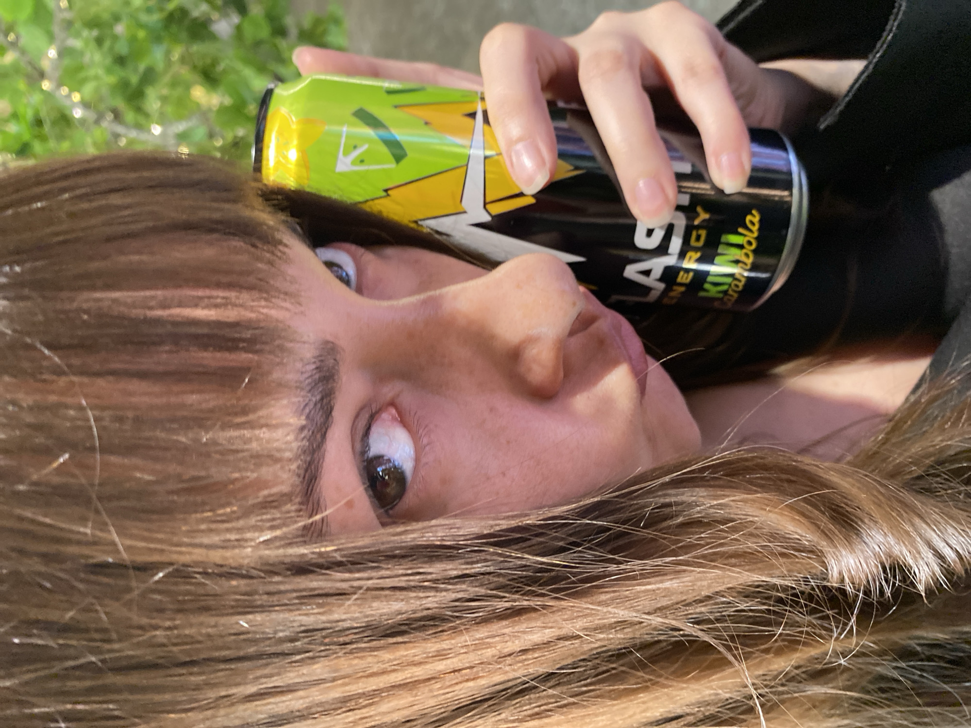 A person lies on their side with their head facing the camera, holding a can of Last Energy Kiwi Carambola drink. The person has brown hair and fair skin. The background is blurred, suggesting an outdoor location with greenery. There is no indication of Votkinsk, Russia in the image.