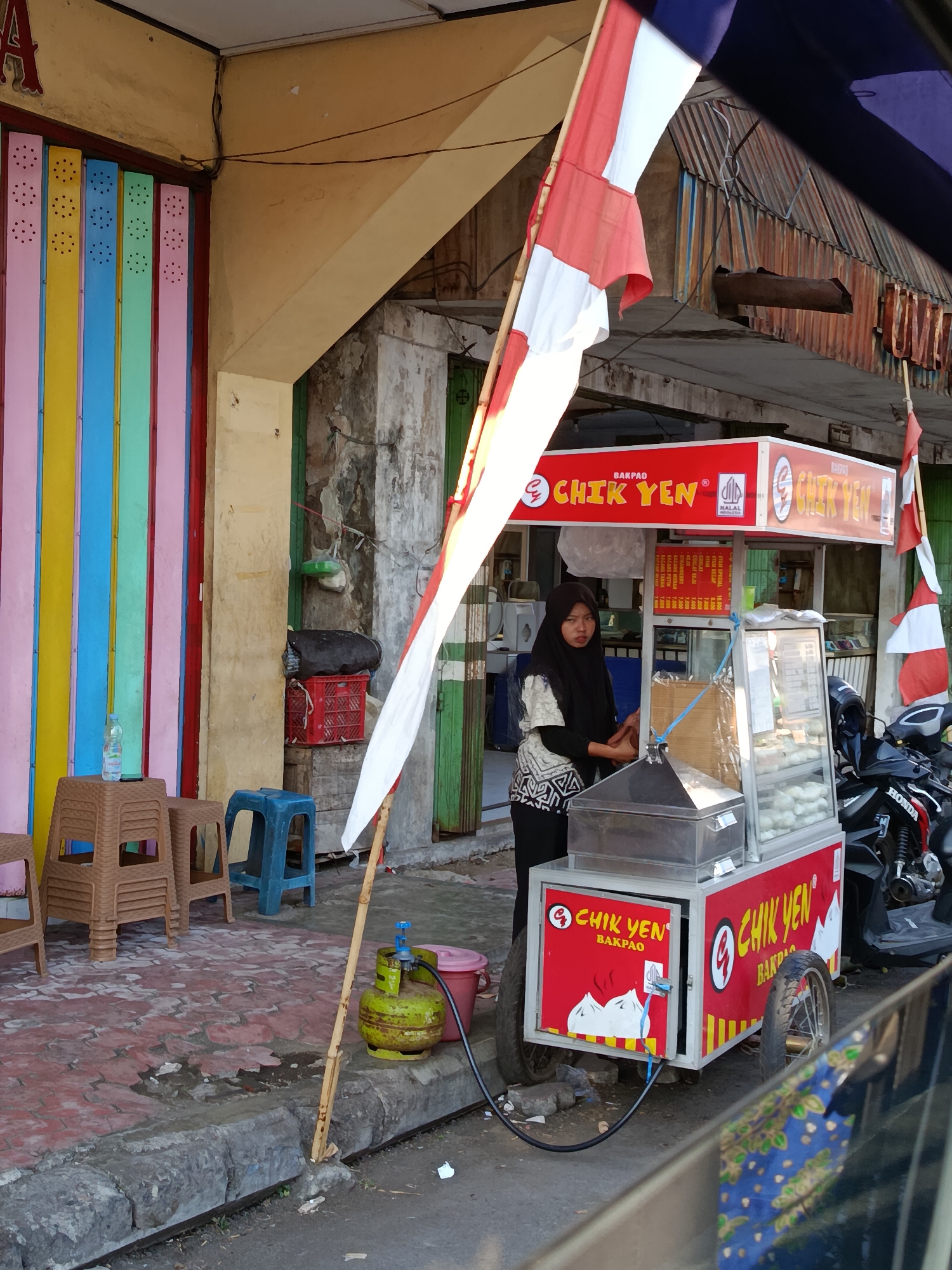 A street vendor stands next to her food cart in Bondowoso, Indonesia. The vendor, a woman wearing a black hijab and a patterned top, is positioned to the left of the cart. The cart is red with "CHIK YEN BAKPAO" branding. Behind her, stacked chairs are visible. To the left of the chairs is a colorful, striped wall. A gas cylinder and a pink bucket are on the ground near the cart. A motorcycle is parked behind the cart. Indonesian flags are prominently displayed, one large flag attached to a pole in front of the cart, and a smaller one on the cart itself. The ground is paved with red tiles, and a concrete curb separates the sidewalk from the street. The backdrop consists of buildings with weathered facades and corrugated metal roofs, suggesting an urban street environment.