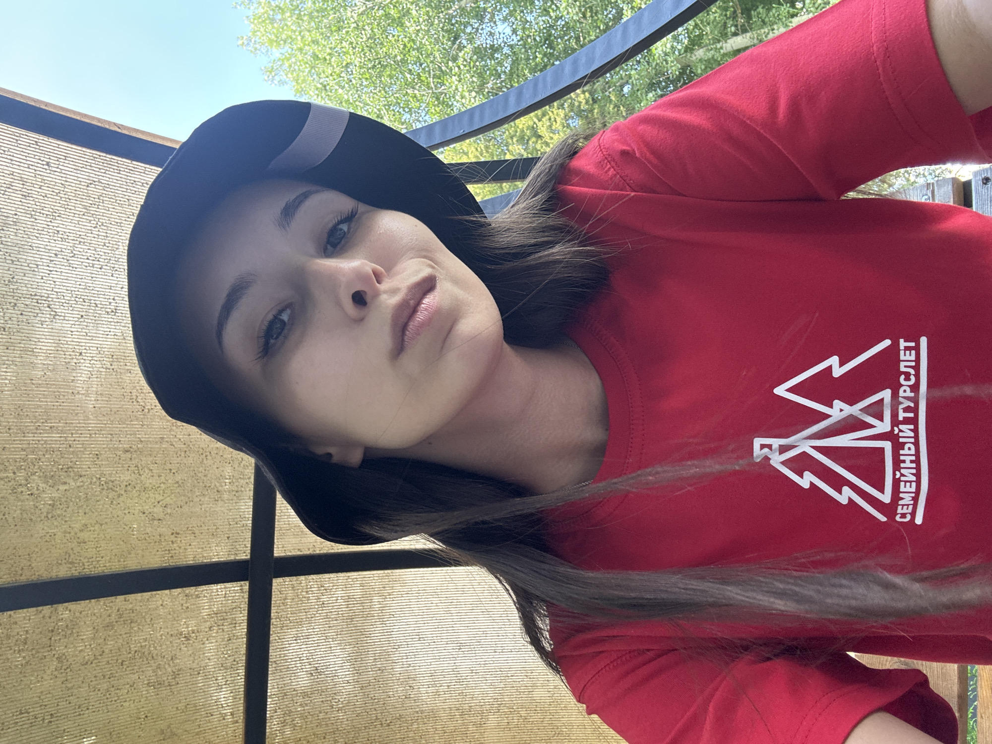 A young woman wearing a black bucket hat and a red t-shirt with a white graphic of trees and the text "СЕМЕЙНЫЙ ТУРСЛЕТ" stands outdoors, looking towards the camera. She has long, dark hair and fair skin. The background consists of a translucent, golden-yellow roof structure above, with green trees and a blue sky visible through it and to the right. A black metal railing is also present on the right side of the image. The scene appears to be in an outdoor recreational area, possibly near a campsite, given the text on the t-shirt which translates to "Family Gathering". The location is Doma 1096 Km, Russia.
