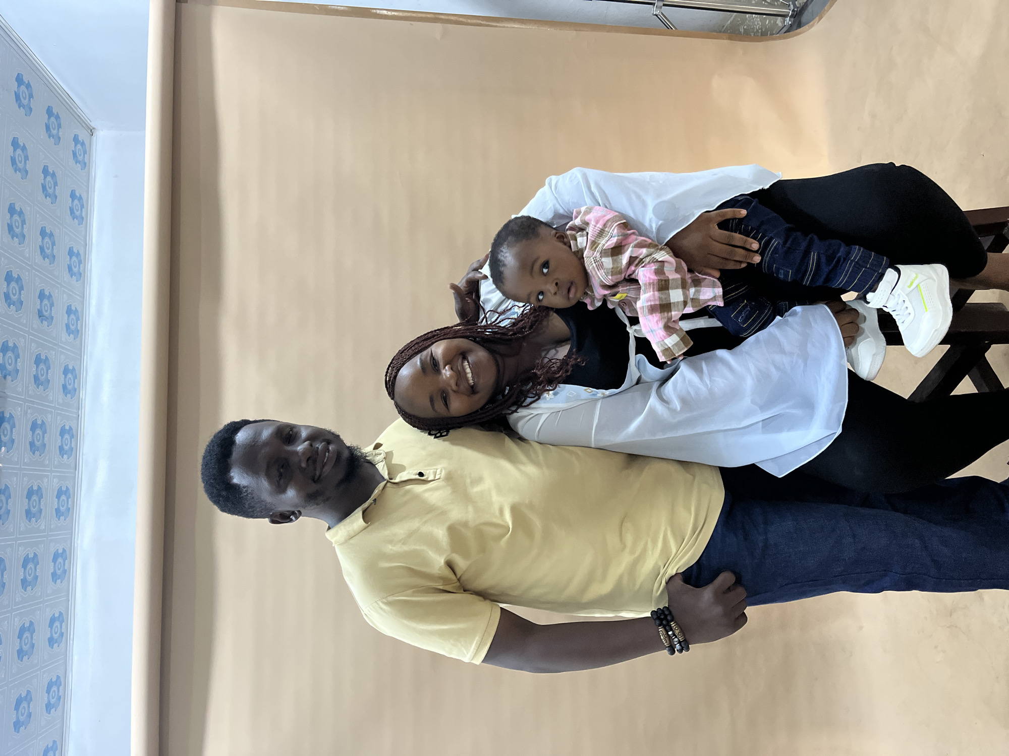 This is a full-body portrait of a family of three, two adults and one infant, posing for a photo against a plain, light brown studio backdrop. The man on the left is wearing a yellow polo shirt and blue jeans, with his left hand in his pocket. He has dark skin and short, dark hair, and is smiling at the camera. The woman in the center is holding an infant on her lap. She has dark skin, braided hair, and is wearing a black top and a white open shirt. She is smiling broadly and looking at the camera. The infant, facing the camera, is wearing a pink and brown checkered shirt and dark blue jeans. The background appears to be a studio setting, with a light brown paper backdrop. Above the backdrop, a ceiling with a repetitive blue and white pattern is visible. The lighting is even and appears to be from studio lights. The scene suggests a casual family portrait session, likely in an indoor studio environment. There is no visible indication of the specific location within the city of Gura Top, Nigeria.
