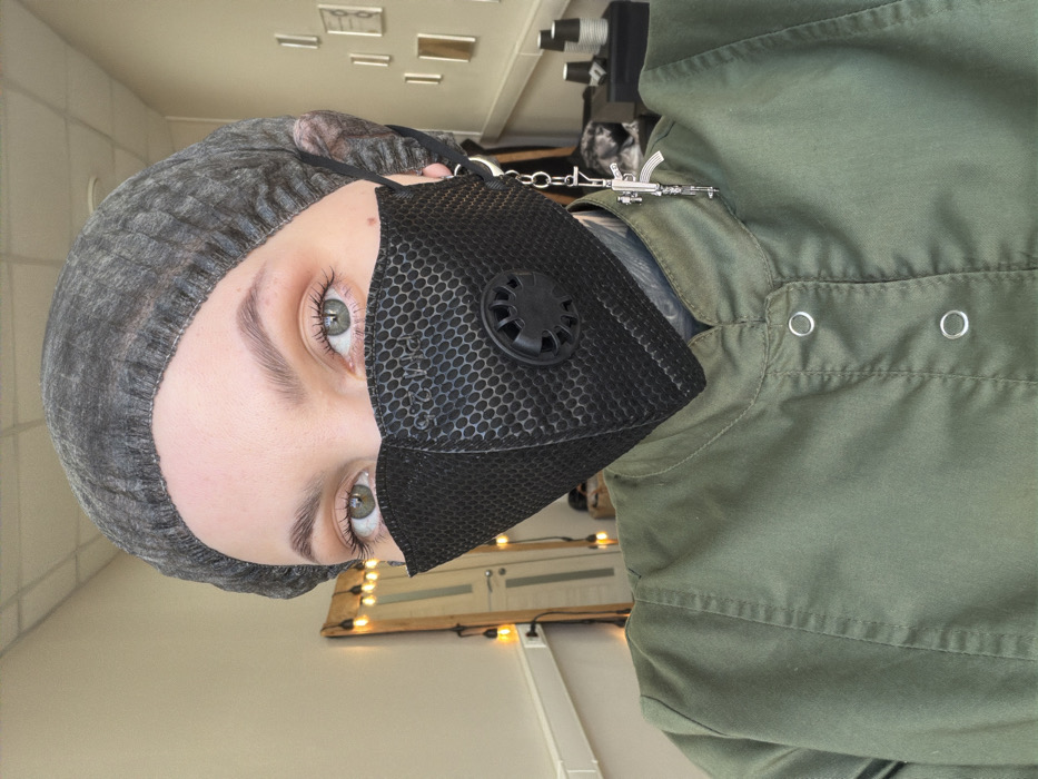 A person wearing a gray head covering and a black face mask with a circular vent is shown. They have light skin, defined eyebrows, and green eyes. They are also wearing a green collared shirt with metal snap closures. A silver chain connects the mask to a miniature machine gun pin on the shirt. The background includes a wall with lights and some visible equipment. The image is vertically oriented.