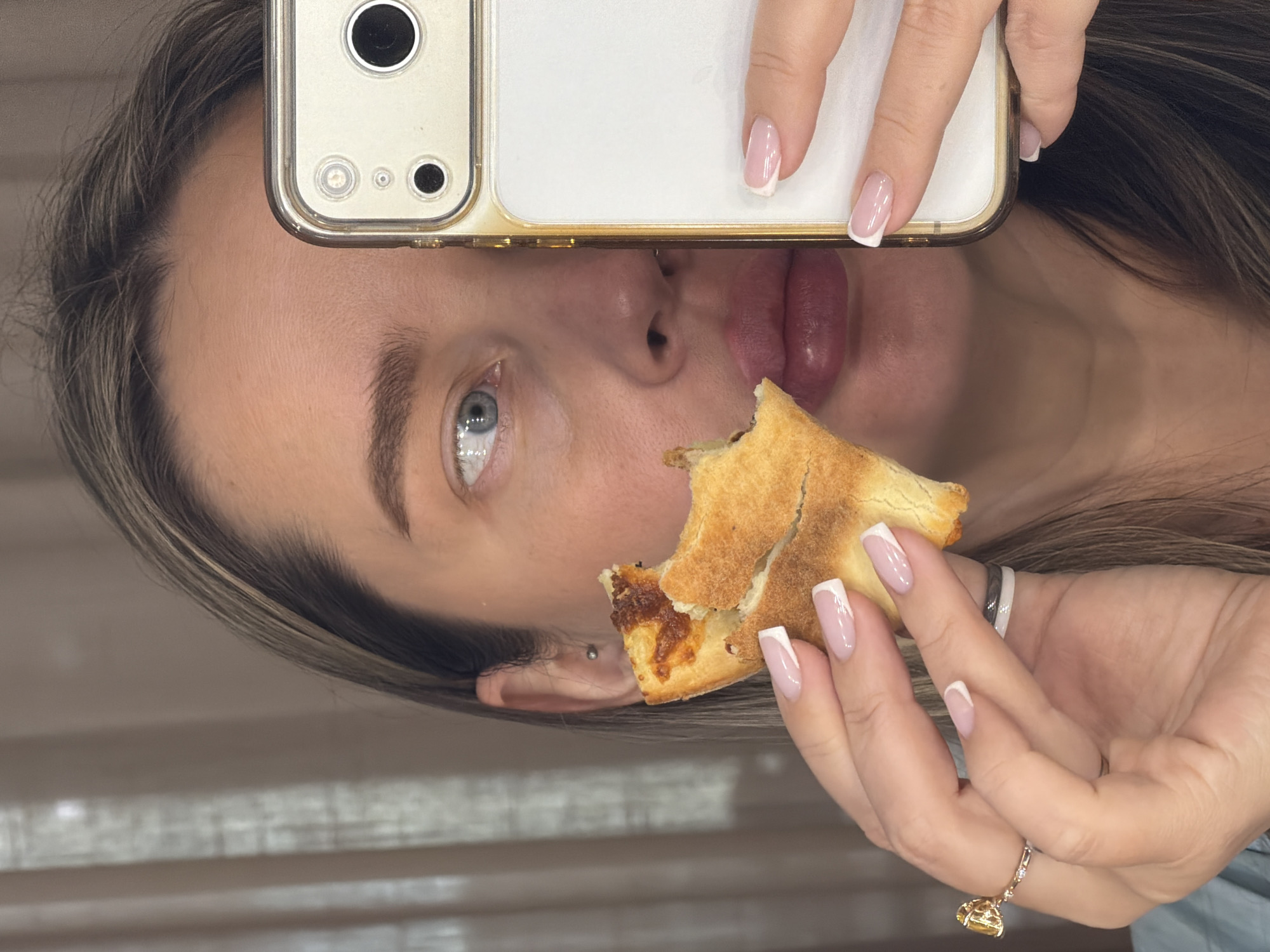 A woman is positioned in front of what appears to be a beige window, holding a partially eaten piece of pizza in her right hand and a white smartphone in her left hand. The phone is positioned such that it covers part of her forehead. The pizza crust is visible, and some of the filling is missing, indicating it has been eaten. The woman has light skin, blue eyes, and brown hair. Her nails are manicured with a French tip. She is wearing rings on her fingers, including one with a dangling gold charm. Due to the lack of contextual details, there is no information that would allow for identification of the location as Moscow, Russia.