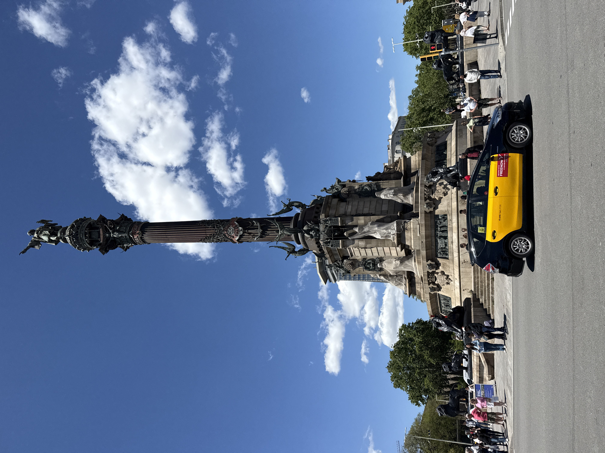 The image depicts the Columbus Monument in Barcelona, Spain, under a blue sky with scattered clouds. The monument, a tall column topped with a statue of Christopher Columbus, is prominently featured. A black and yellow Barcelona taxi cab is parked on the street next to the monument. Pedestrians are seen walking around the base of the monument. Trees line the street behind the monument.
