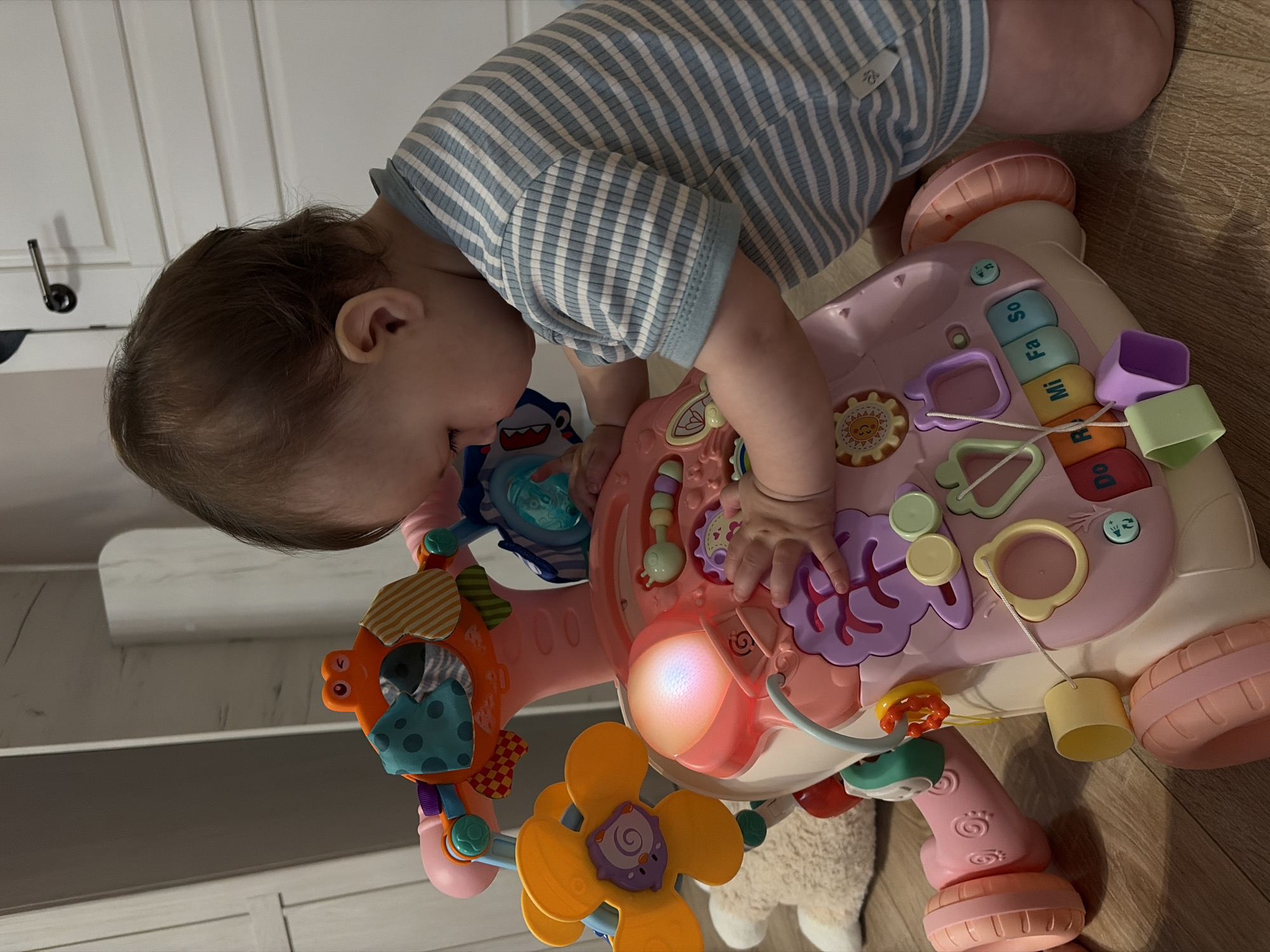 A child is interacting with a pink and white interactive toy. The child is wearing a blue and white striped outfit. The toy features various attachments and activities, including a light-up element, musical keys, and dangling objects. The scene appears to be indoors, with a wooden floor and a glimpse of what looks like a white cabinet or wall in the background. The location is not specified.