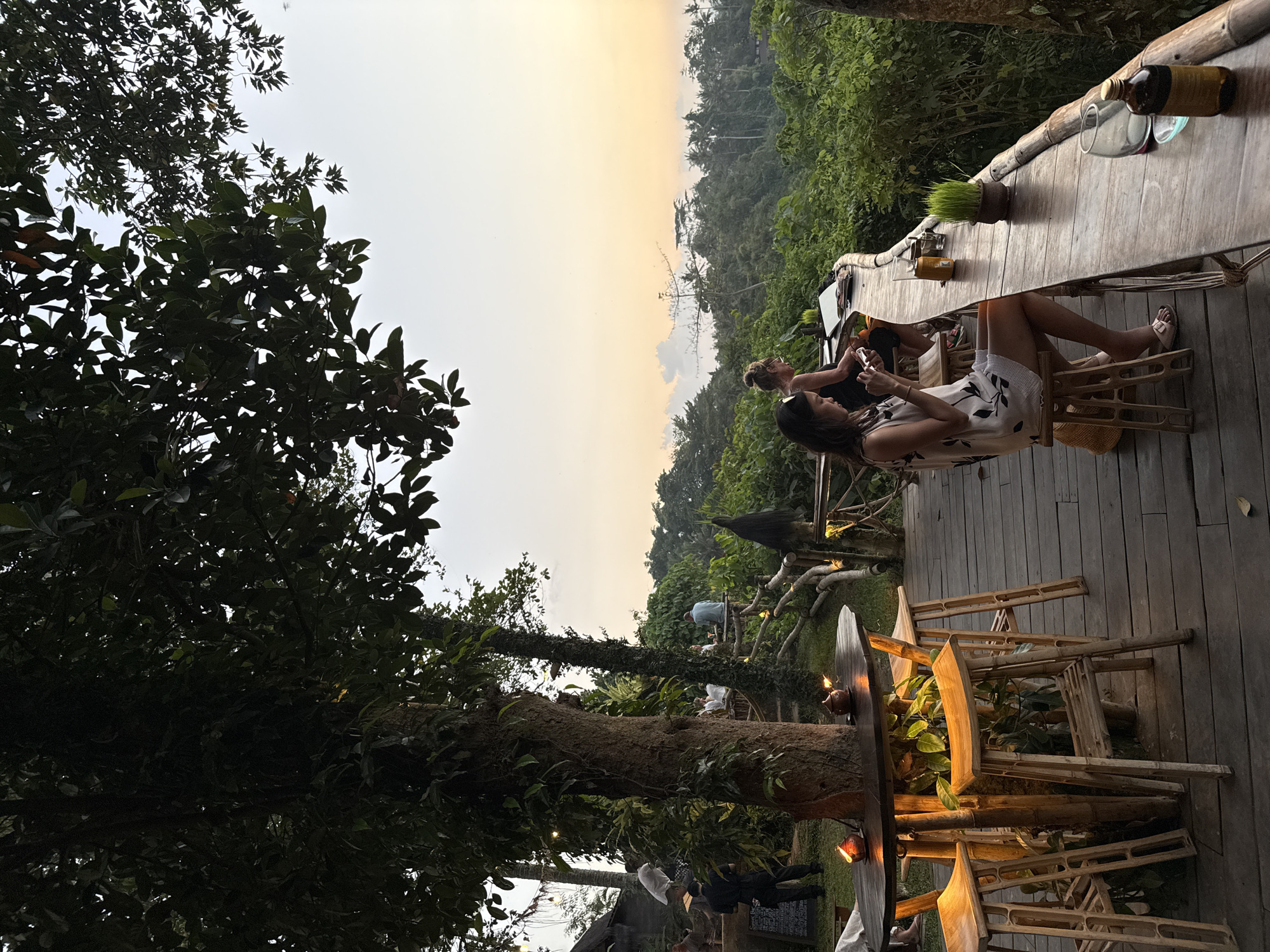 An outdoor cafe in Ubud District, Indonesia, features a wooden deck overlooking a lush, green jungle as the sun sets, casting a warm glow across the sky. On the deck, two women are seated at a long wooden bar. The woman in the foreground, wearing a white dress with a black floral pattern, is looking at her phone. Behind her, another woman, wearing a black top, is also seated at the bar.
In the foreground, a round wooden table is surrounded by three bamboo stools. Two candles in small terracotta holders provide illumination. The cafe is situated amidst large trees with dense foliage, some of which overhang the deck, creating a natural canopy. In the background, more trees and the jungle canopy stretch out to the horizon. Several individuals are visible in the distance, also seated at tables on a raised wooden platform. The overall atmosphere suggests a relaxed evening dining or socializing experience in a tropical setting.