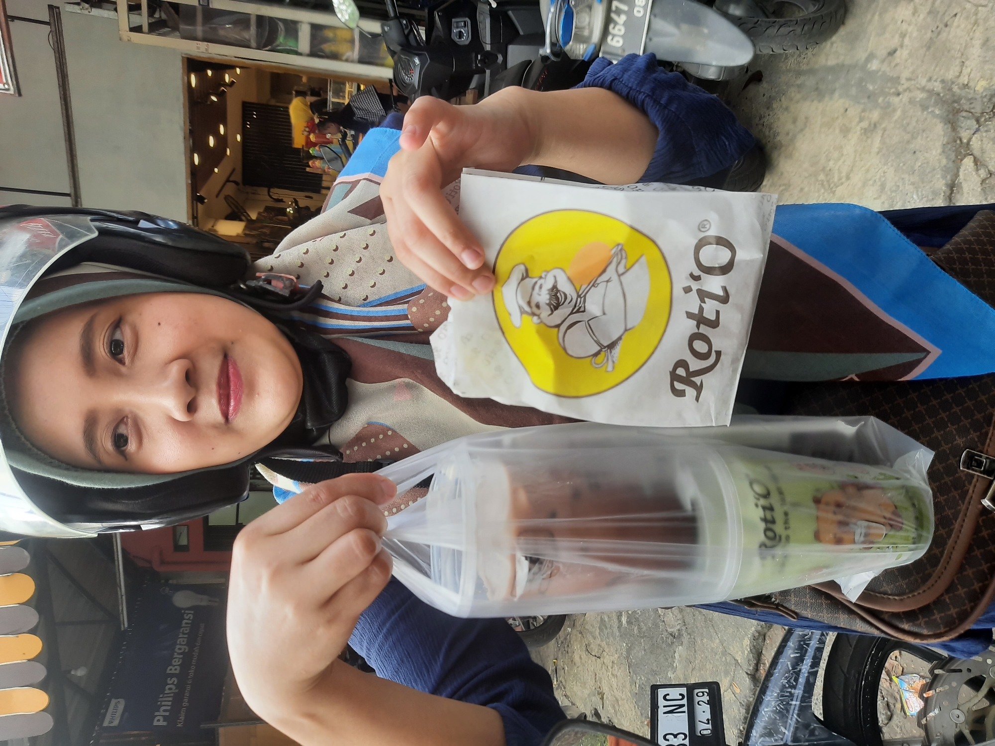 In a street scene in Bandung City, Indonesia, a woman wearing a hijab and motorcycle helmet is holding two takeaway items. She is positioned in the foreground, facing the camera. In her right hand, she holds a clear plastic bag containing a beverage with a brown liquid and ice. Her left hand supports a paper bag with a yellow circular logo featuring a cartoon chef and the text "Roti'O". The background shows a street with parked motorcycles, some shop fronts, and a banner that appears to be for "Philips Bergaransi". The overall environment suggests an urban setting, possibly a commercial area.