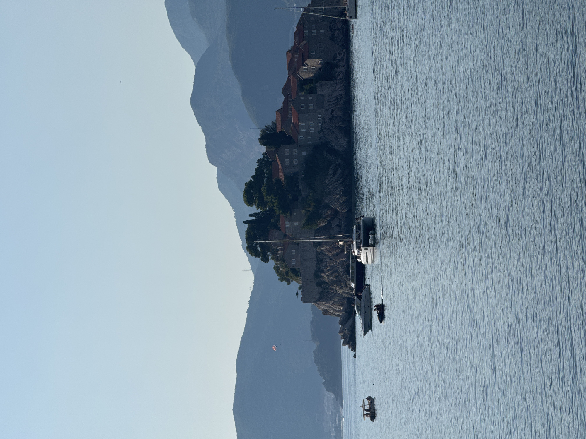 The image depicts the Sveti Stefan island resort in Montenegro. The island is a fortified village built on a rocky promontory, featuring stone buildings with red-tiled roofs. It is surrounded by the clear blue waters of the Adriatic Sea. Several boats are visible in the foreground, including a small passenger boat, a jet ski with two individuals, and at least two sailboats. A parasailer can be seen in the sky above the island. In the background, lush green hills and mountains rise, creating a scenic backdrop. The sky is a pale, clear blue, suggesting a sunny day. The overall scene conveys a sense of leisure and natural beauty.