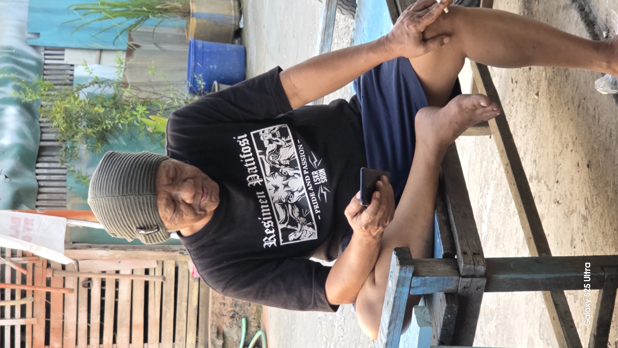 In Juwana, Indonesia, an elderly person wearing a green beanie and a black t-shirt with the text "Resimen Patifosi - Pride and Passion" is seated on a rustic wooden bench. The individual is looking down at a smartphone held in their left hand. Their right hand rests on their knee, holding a cigarette. The background consists of a corrugated metal fence and some greenery, suggesting an outdoor, possibly residential, setting. The bench appears weathered and is painted blue in some areas. The ground is paved with concrete and dirt. The lighting indicates it is daytime.