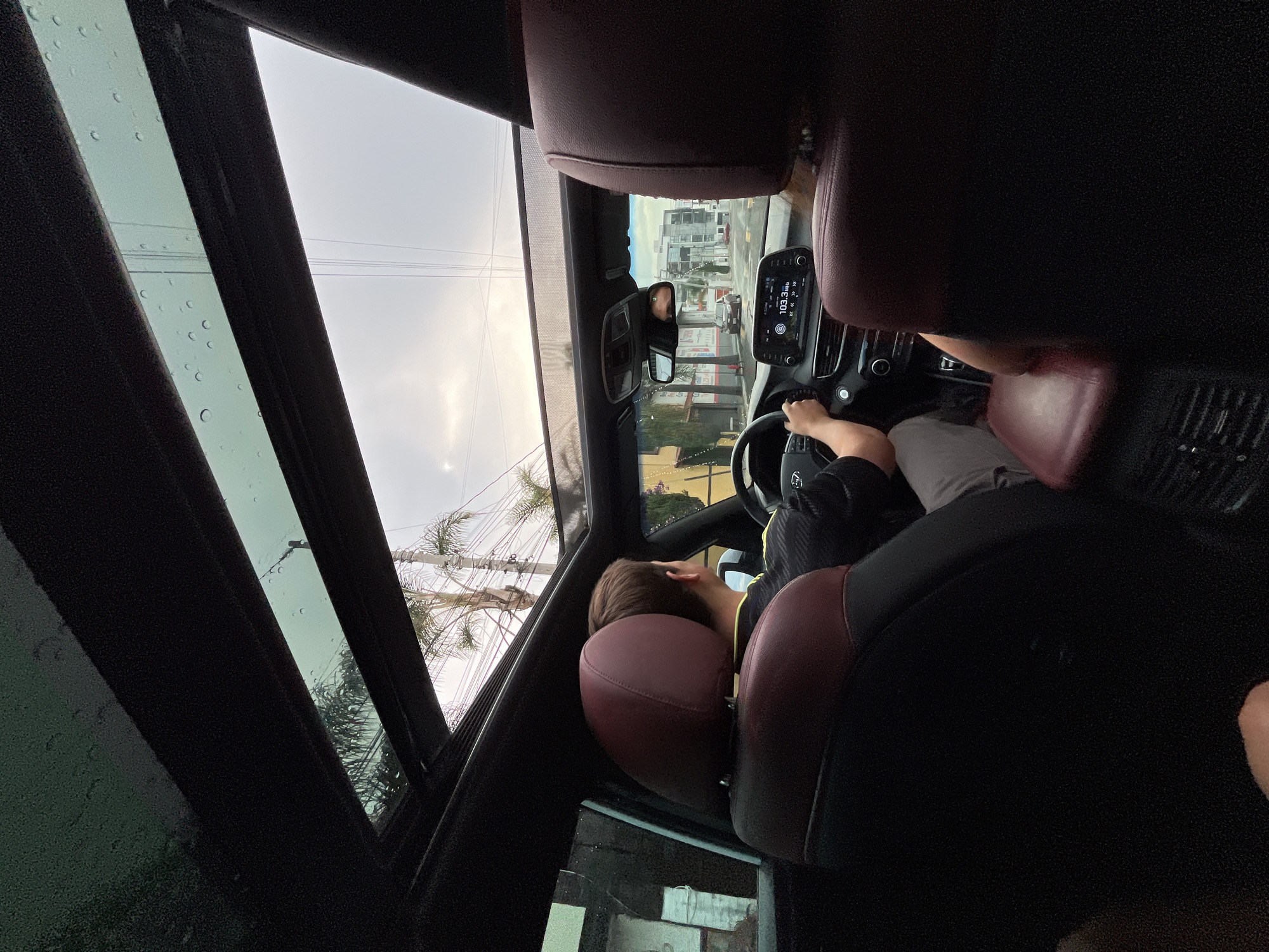 The image depicts the interior of a car, possibly a SUV, with a partial view of the driver and the passenger seat behind the driver. The driver is holding the steering wheel, with their head resting on the seat. The driver appears to be wearing a black shirt. The interior features burgundy or dark red leather seats. The dashboard is visible with a digital display showing a radio frequency of 103.3 MHz. Outside, the scene shows a cloudy sky and some buildings, palm trees and utility poles. This location could possibly be in San Bernardino Tlaxcalancingo, Mexico. There are water droplets visible on the glass roof.