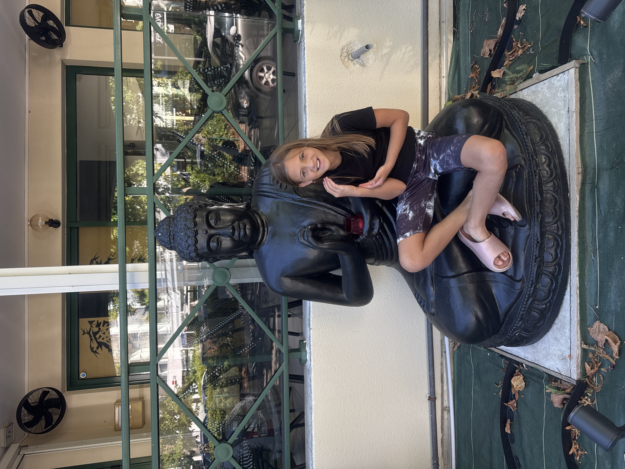 A young girl with blonde hair and blue eyes is sitting on a large black statue of Buddha in Divonne-les-Bains, France. The girl is wearing a black t-shirt and patterned shorts, with pink sandals on her feet. She is smiling and looking at the camera. The statue of Buddha is seated and is dark black, with its hands in a meditative pose. In front of the statue, there is a small red candle. The background shows a building with large windows, a green metal railing, and trees outside. There are also two black fans mounted on the wall of the building. The ground is covered with a green mat and scattered autumn leaves.
