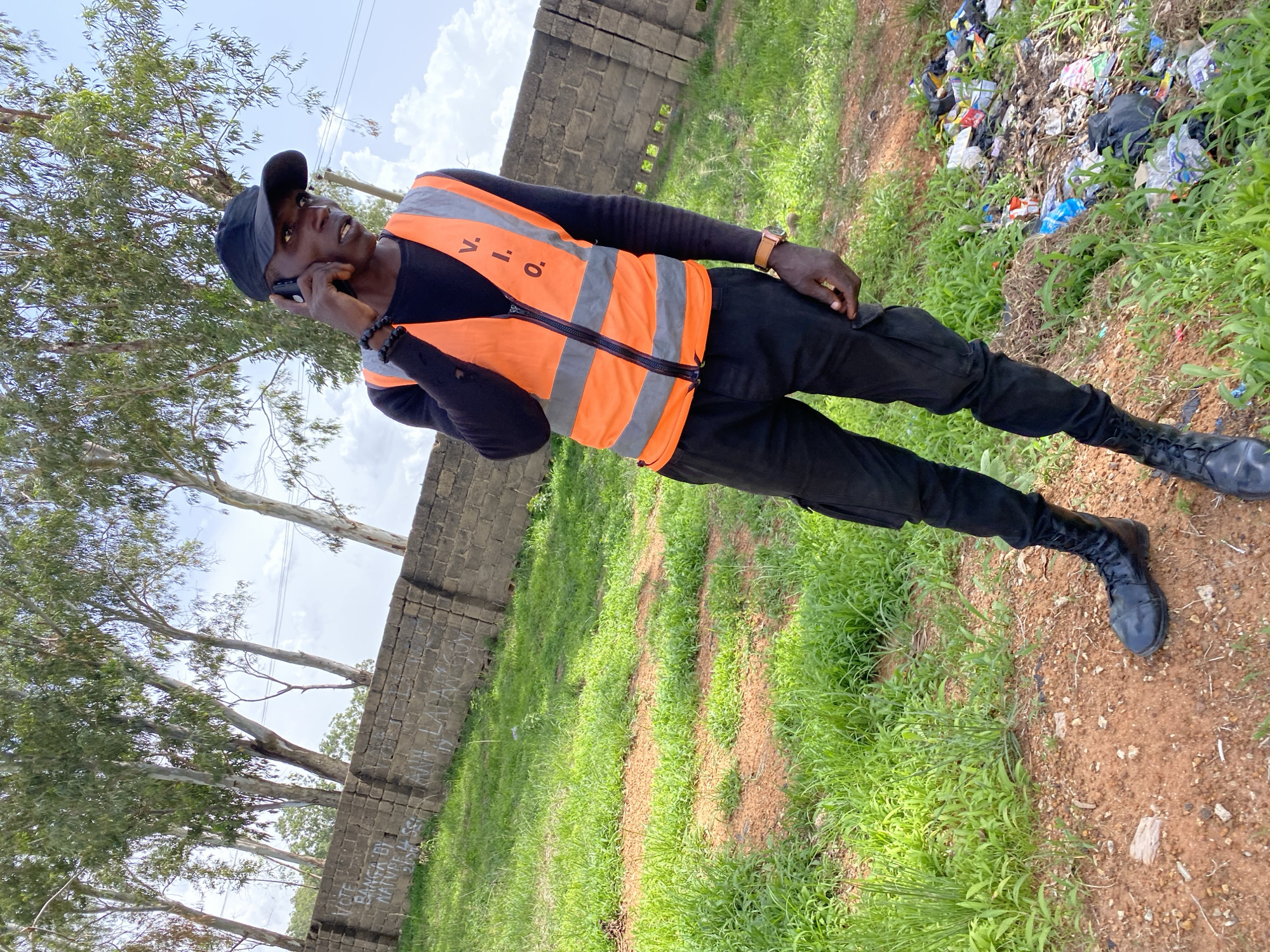 The image shows a full-body shot of a Black man standing outdoors, wearing a black cap, black long-sleeved shirt, an orange high-visibility vest with grey reflective strips, black cargo pants, and black boots. He is holding a black mobile phone to his left ear, engaged in a conversation. The man has dark skin and is wearing a brown wristwatch on his right wrist. He also has a black beaded bracelet on his left wrist.
The setting appears to be a somewhat rural or undeveloped area. To the left of the man, there is a wall constructed from grey bricks, with some graffiti visible on it. Behind the wall, tall trees with green foliage are visible against a bright, cloudy sky, suggesting it is daytime. To the right of the man, there is a patch of dry, reddish-brown earth covered with green grass and scattered debris, including what looks like trash. The ground is uneven.
The man's expression is difficult to determine clearly due to the angle and resolution, but he seems focused on his phone call. The overall atmosphere suggests a casual moment during a potentially work-related activity, given the high-visibility vest. The location context provided is Bukuru, Nigeria. No other specific event or action is discernable beyond the man's phone call.