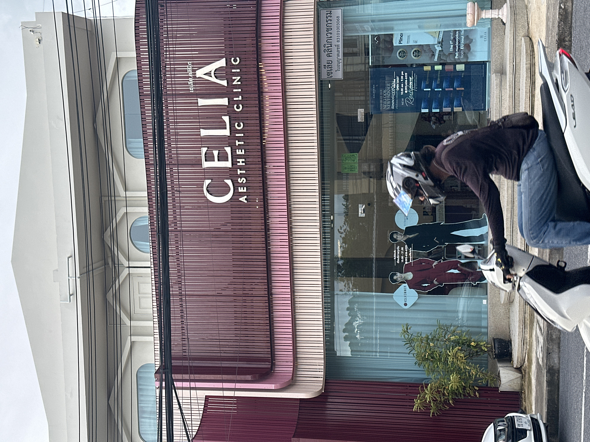 This is a vertical, eye-level shot of the exterior of a clinic in Phuket City Municipality, Thailand. The clinic, named "CELIA AESTHETIC CLINIC", is prominently displayed on the building facade with large, silver lettering. The building features a modern design with a combination of dark red and lighter pink horizontal slats, interspersed with vertical white lines. Large windows reveal the interior, showcasing displays related to cosmetic treatments.
A person wearing a dark jacket, blue jeans, and a white helmet is partially visible, leaning over a white scooter parked on the sidewalk. The person appears to be interacting with something on the scooter, perhaps adjusting it or checking something. The scooter's handlebars and part of its front are visible, along with the rear of another white vehicle further down the street.
Signs on the clinic's windows provide information about its services, opening hours, and contact details. One sign reads "GALDERMA INTRODUCE OUR NEW LOOK NEW PACKAGE" and lists operating hours as "OPEN DAILY 10.00 am - 9.00 pm" with a phone number. Another sign is in Thai and mentions "เซเลีย คลินิกเวชกรรม" (Celiah Clinic of Medicine) and a license number.
A small green tree in a pot stands on the sidewalk in front of the clinic. The sky appears overcast, suggesting a cloudy day, and the scene suggests a typical daytime urban environment. The overall impression is that of a busy street scene in a commercial area.