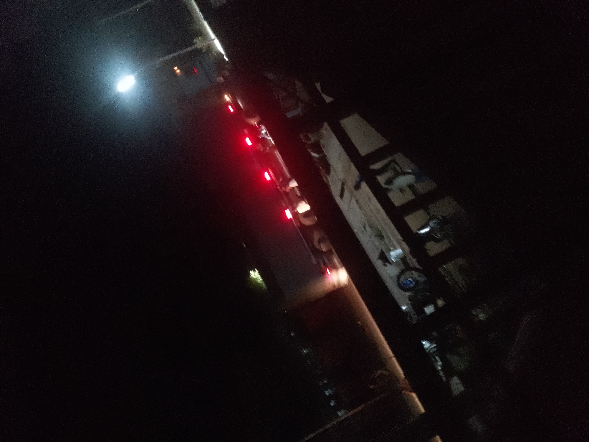 A long vehicle, likely a truck or bus, is parked on an unpaved road at night in Jalingo, Nigeria. The vehicle has several red lights illuminated along its length. A bright white light is visible in the upper left portion of the image, possibly from a streetlamp or another vehicle. The surrounding environment is dark, with some indistinct structures and foliage visible. There are no people clearly identifiable in the image.