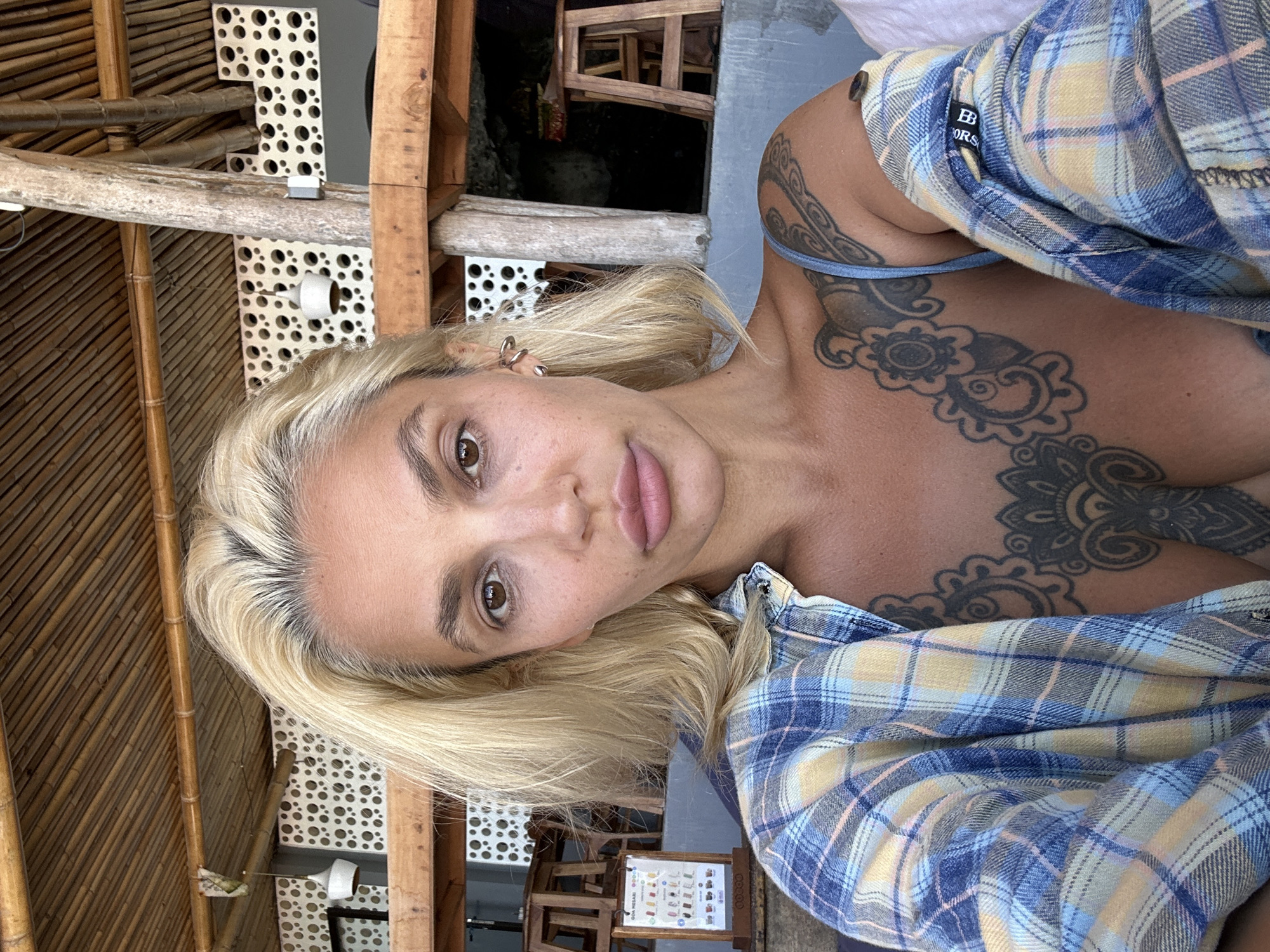 An adult female is depicted in a close-up, self-taken portrait within an open-air or semi-open-air establishment. She has blonde hair with visible dark roots, brown eyes, and full lips. Dark circles are present under her eyes. Multiple small, curved, metallic ear cuffs are visible on her right ear. She has extensive black ink tattoos, including an intricate floral or mandala-style design spanning her sternum and upper chest, and a swirling, organic pattern on her left shoulder and upper arm. She is wearing a light blue strap top or bralette beneath an unbuttoned, long-sleeved plaid shirt with a pattern of blue, yellow, and thin pink lines.
The background features a ceiling constructed from parallel bamboo reeds. Supporting structures include rough-hewn wooden beams and a prominent, light-colored tree trunk pillar. Beyond these, white partition walls with numerous circular cutouts of varying sizes are visible. A white, bell-shaped pendant light hangs on the left. To the left, a small, wooden A-frame sign displays a menu with "GULAI MASABI" at the top, indicating a food and beverage establishment. In the deeper background, wooden stools are partially visible, suggesting a dining area, along with a dark, natural cave-like recess behind the large pillar. The architectural style and the "Gulai Masabi" menu sign are consistent with establishments found in Kuta Selatan, Indonesia.