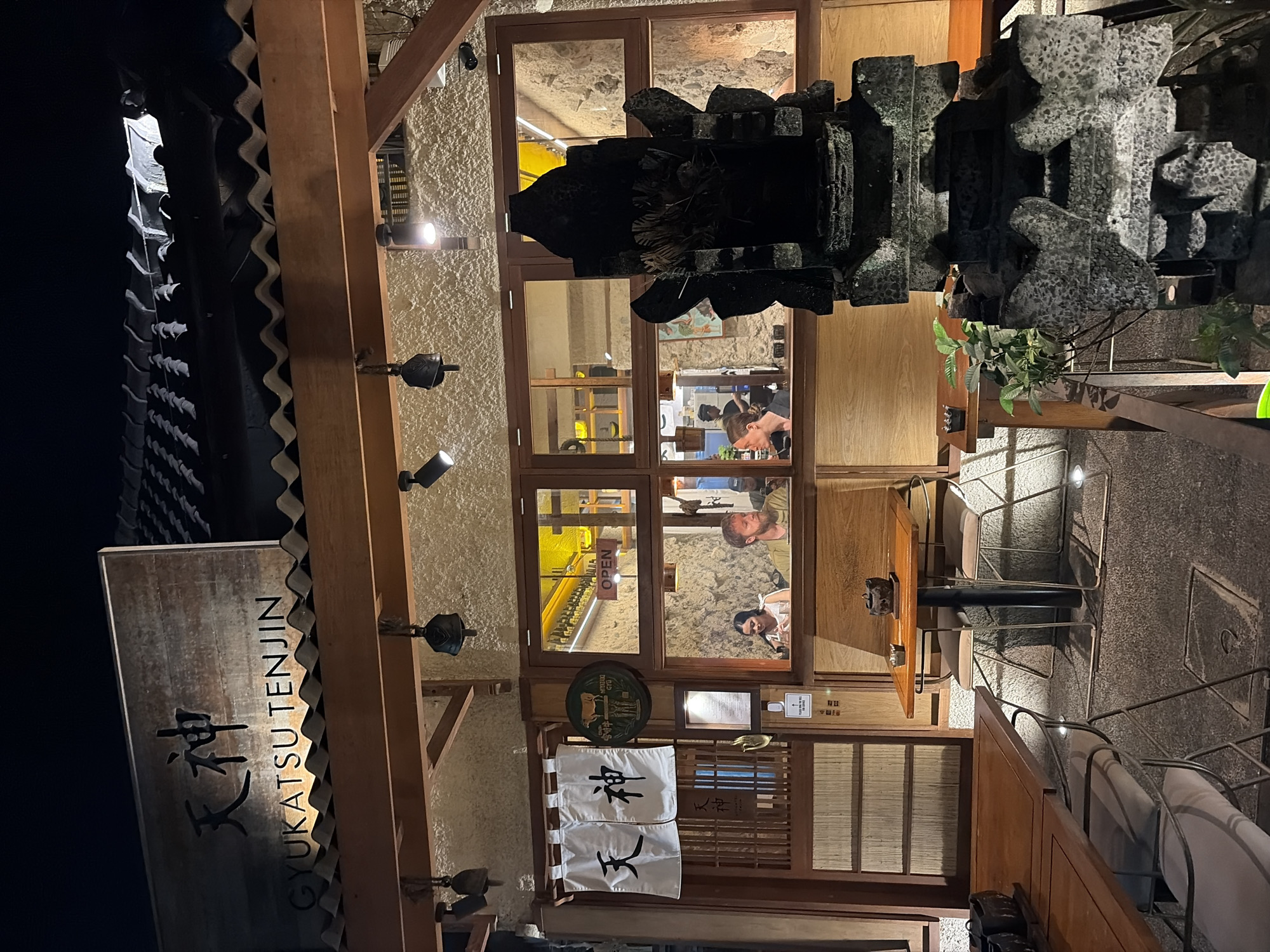 The image shows the exterior of a Japanese restaurant at night in the Ubud District, Indonesia. The restaurant has a traditional Japanese facade with wooden elements and a tiled roof. A sign above the entrance reads "GYUKATSU TENJIN" in English and features Japanese characters. Inside the restaurant, visible through large windows, are patrons seated at tables. One table near the window has two people, a man and a woman, who appear to be dining. Another area of the restaurant shows what looks like a kitchen or bar area with shelves lined with bottles. A traditional stone sculpture stands in the foreground, adding to the ambiance. The lighting is warm, coming from interior lights and spotlights illuminating the facade. The ground outside is paved with gravel.
