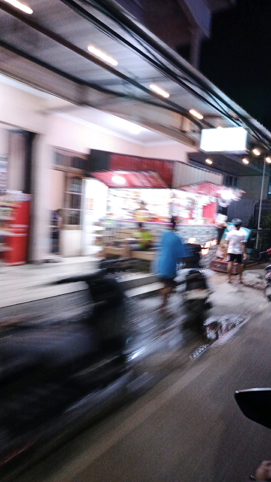 The image depicts a blurred nighttime scene on a street in Surakarta, Indonesia, likely near a row of shops or food stalls. The motion blur suggests movement, possibly from a vehicle or a panning camera.
In the background, a building with a pink facade is visible, featuring a red awning and illuminated signs, indicating commercial activity. Several people are present, though their features are indistinct due to the blur. One individual in the mid-ground appears to be wearing a bright blue garment. Another person, further to the right, is wearing a white shirt and dark shorts.
In the foreground and mid-ground, several motorcycles are visible, their forms elongated by the motion blur. The street surface appears wet, reflecting some of the ambient light and suggesting recent rain. Streetlights and overhead lighting contribute to the overall illumination of the scene. The overall impression is a dynamic, bustling urban environment at night.